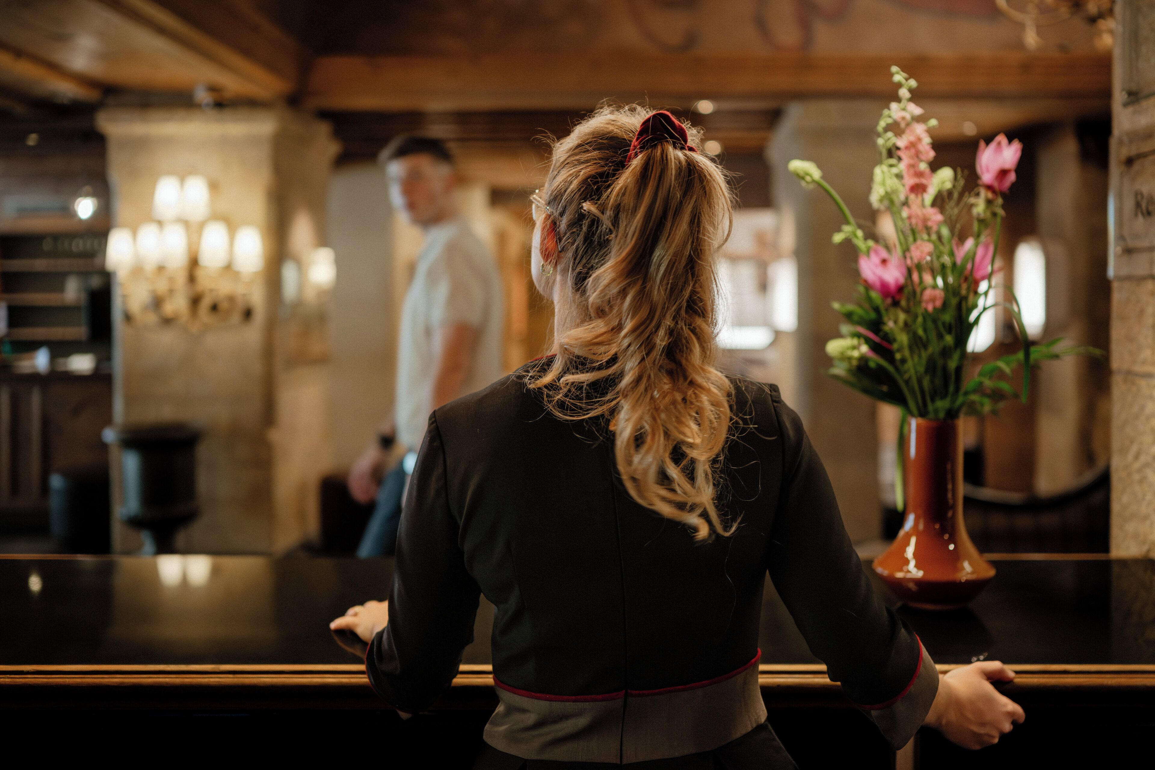 Una persona está de pie en la recepción de un hotel, con un jarrón de flores y una araña en el fondo.