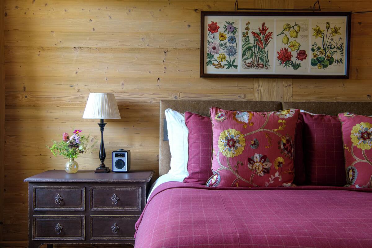 A bedroom with a wooden wall, a framed botanical print, a bedside table with a lamp, a radio, and a vase with flowers.