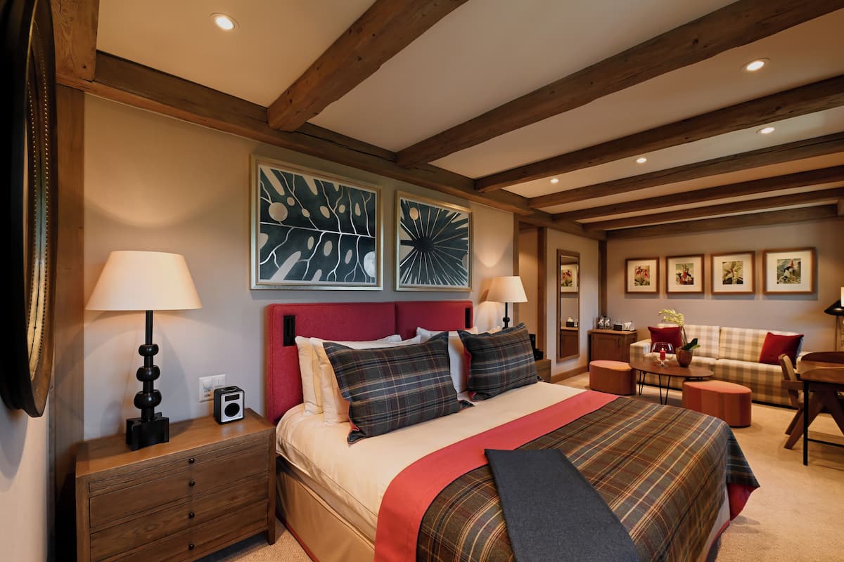 A hotel room with a large bed, a red headboard, two wall-mounted lamps, a wooden nightstand, a red throw blanket, and a checkered sofa.