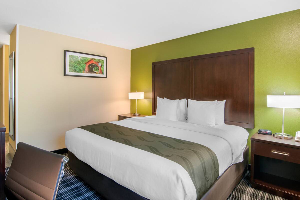 Una habitación de hotel con una cama grande, una silla, una mesita de noche con una lámpara y un cuadro en la pared.