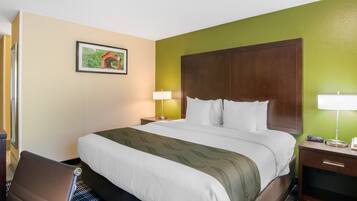 Una habitación de hotel con una cama grande, una silla, una mesita de noche con una lámpara y un cuadro en la pared.