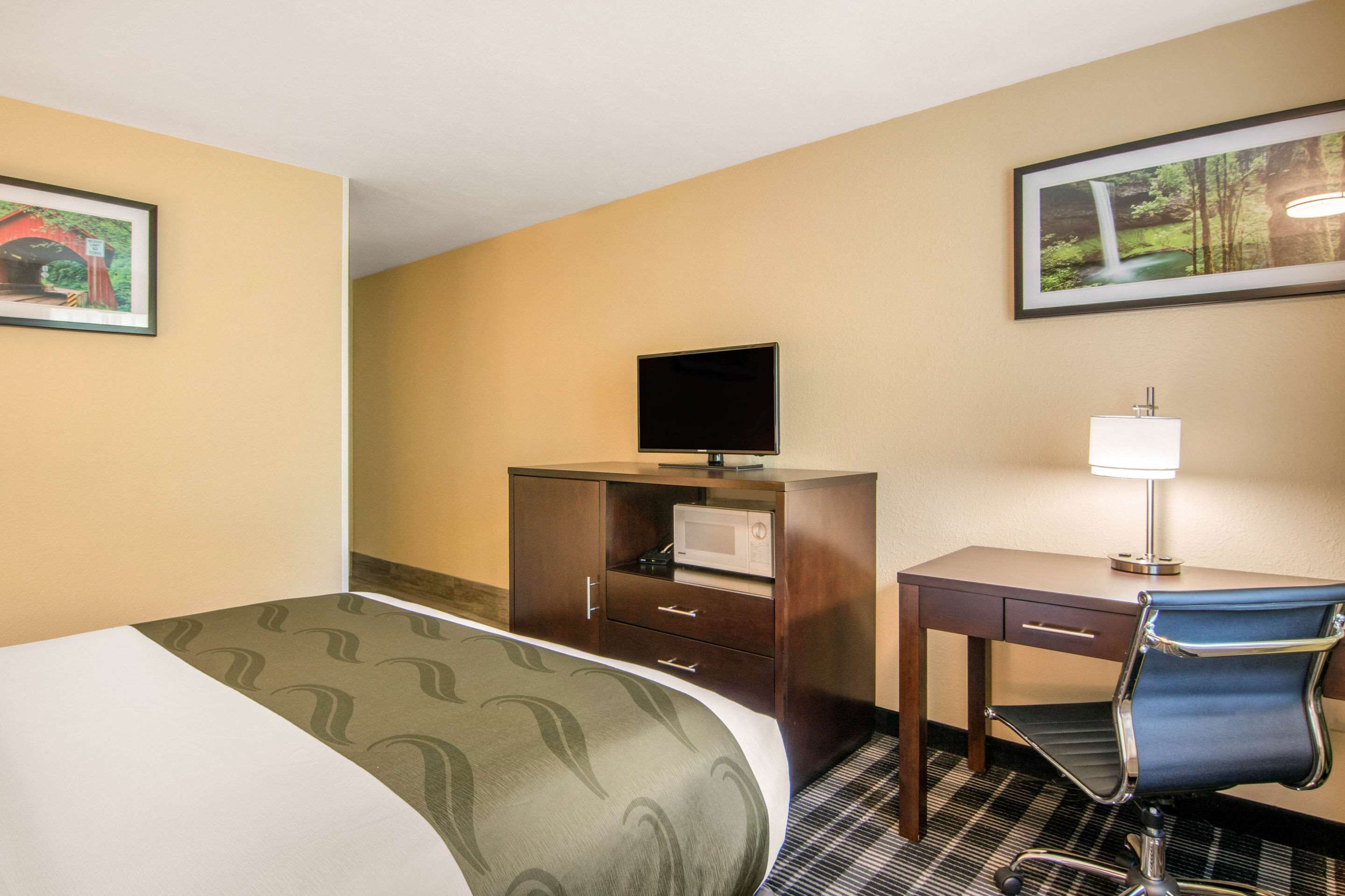 Une chambre d’hôtel équipée d’un lit, d’un bureau, d’une chaise, d’une télévision, d’un four à micro-ondes et de deux tableaux encadrés accrochés aux murs.