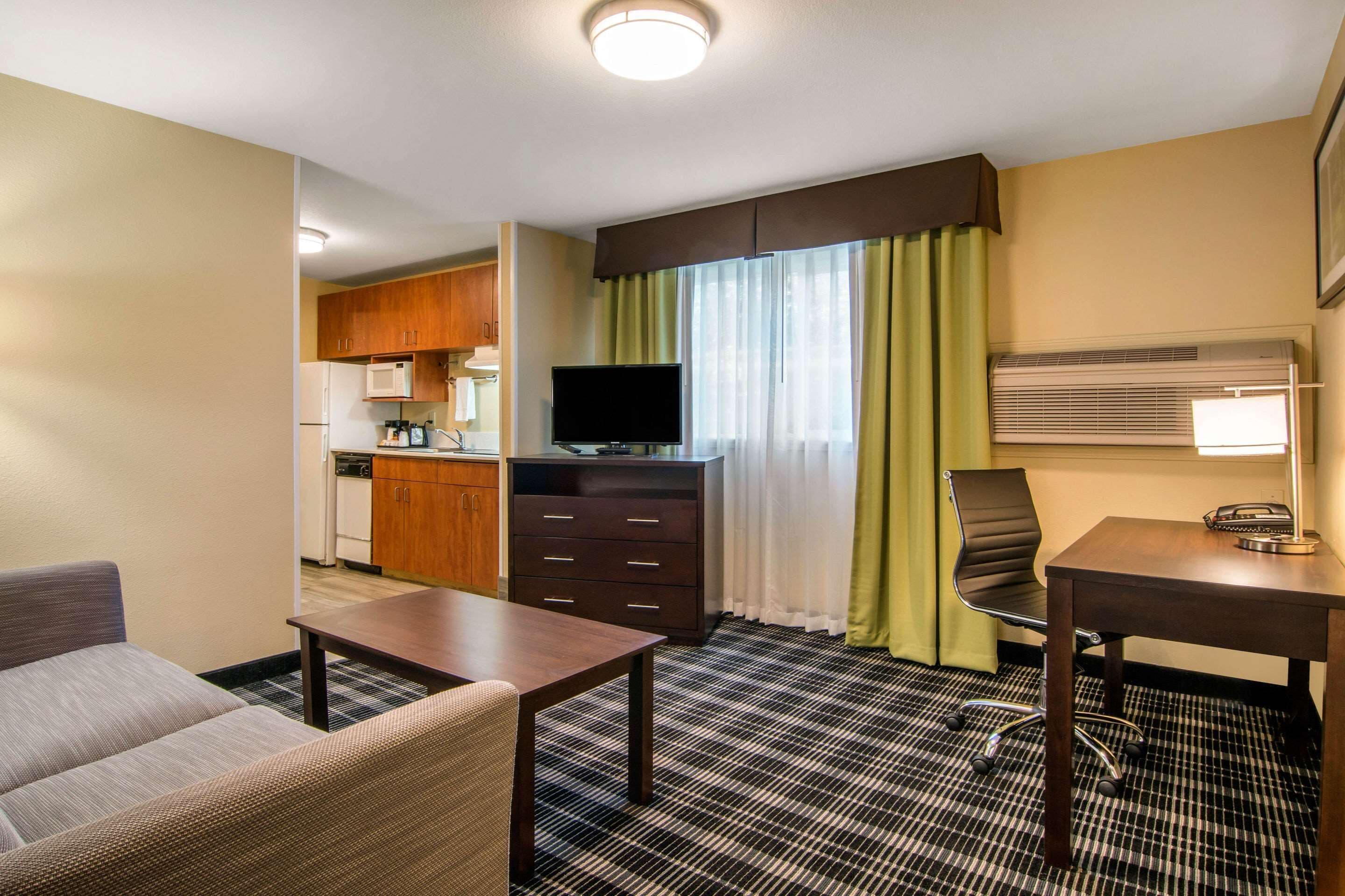 A hotel room with a kitchenette, a television, a desk with a lamp, a chair, and a sofa.