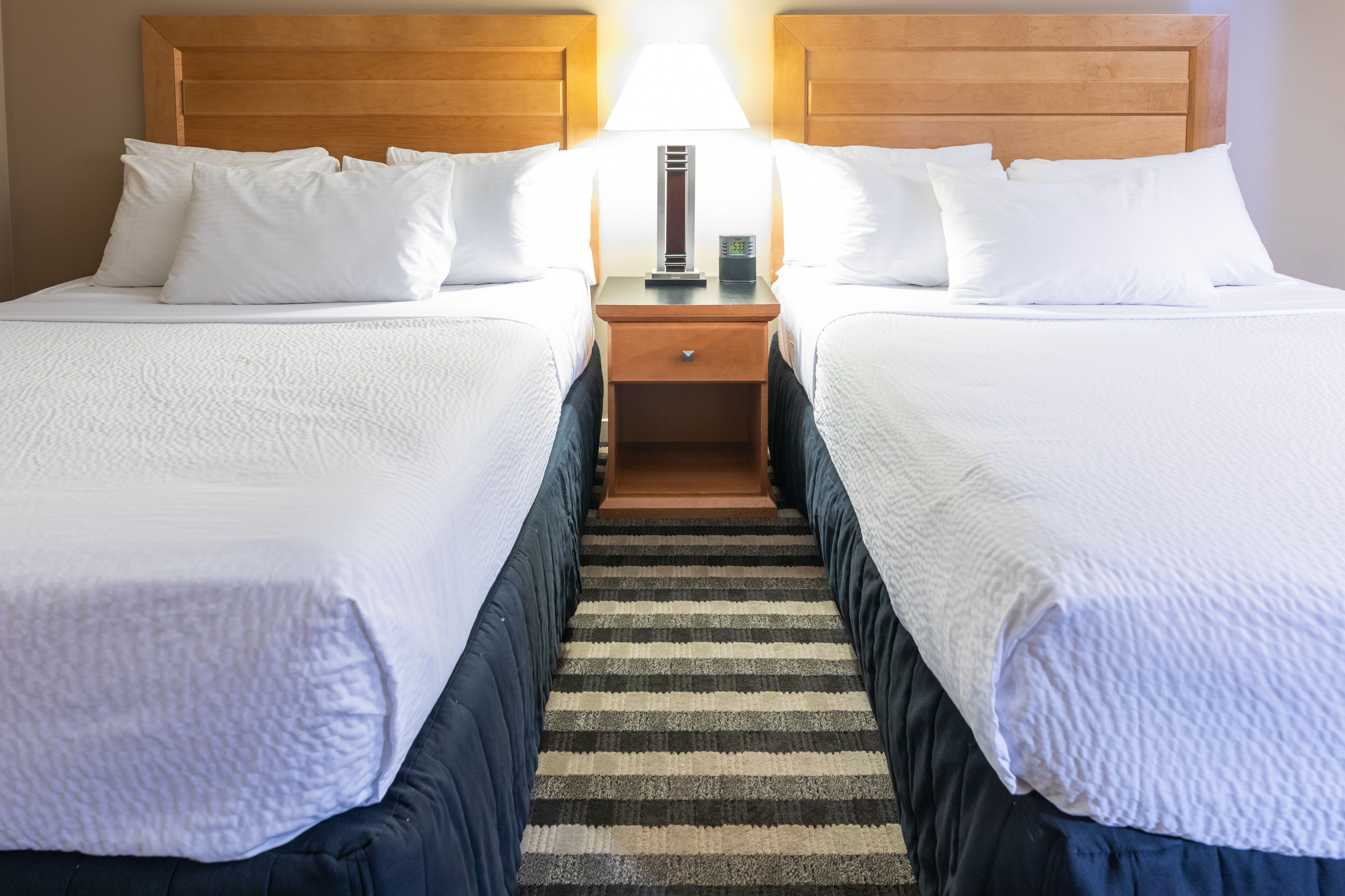 A hotel room with two beds, a nightstand with a lamp, and a striped rug.