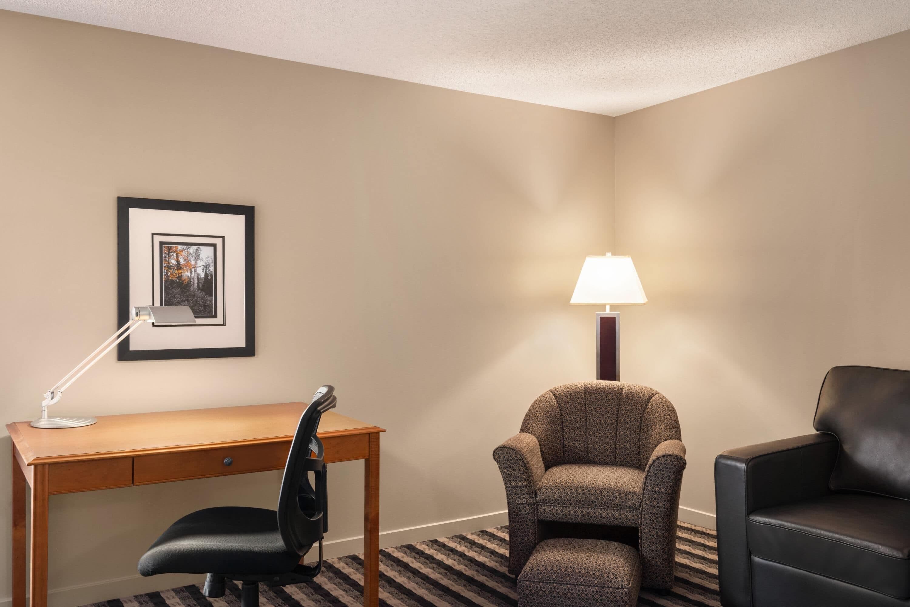 A hotel room with a wooden desk, a black office chair, a patterned armchair, a black sofa, a framed picture on the wall, and a standing lamp.