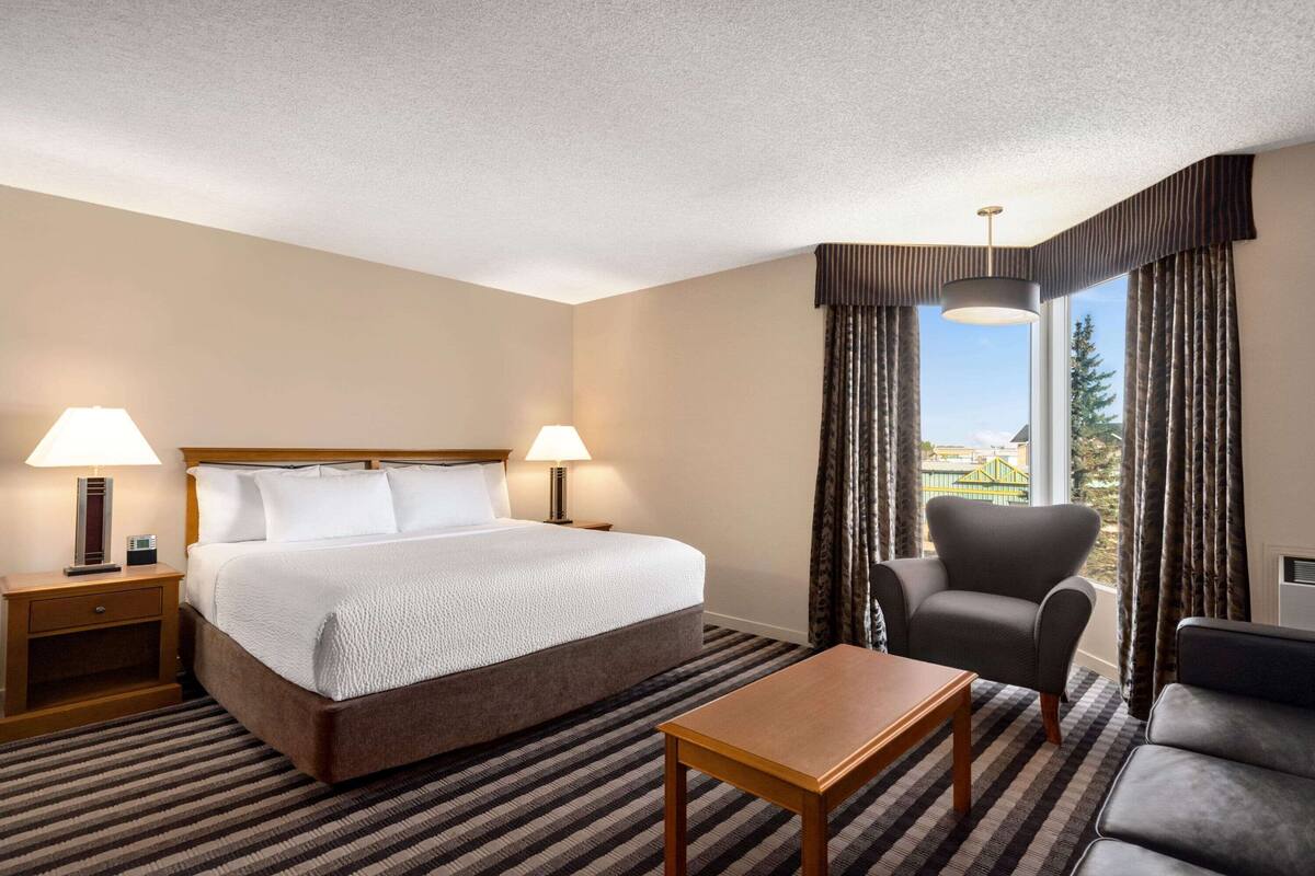 A hotel room with a bed, two bedside lamps, a chair, a table, and a view of the outside.