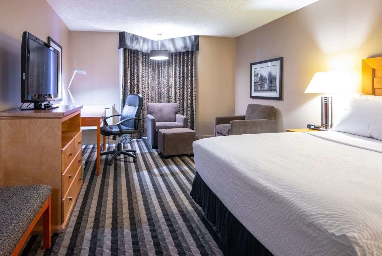 A hotel room with a bed, a desk with a computer, two armchairs, a chair, a TV, and a lamp.