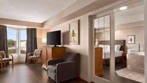 A hotel room with a television, a sofa, a bed, a bathroom with a bathtub, and a large window with curtains. - Royal Hotel West Edmonton, Trademark Collection by Wyndham (Edmonton)