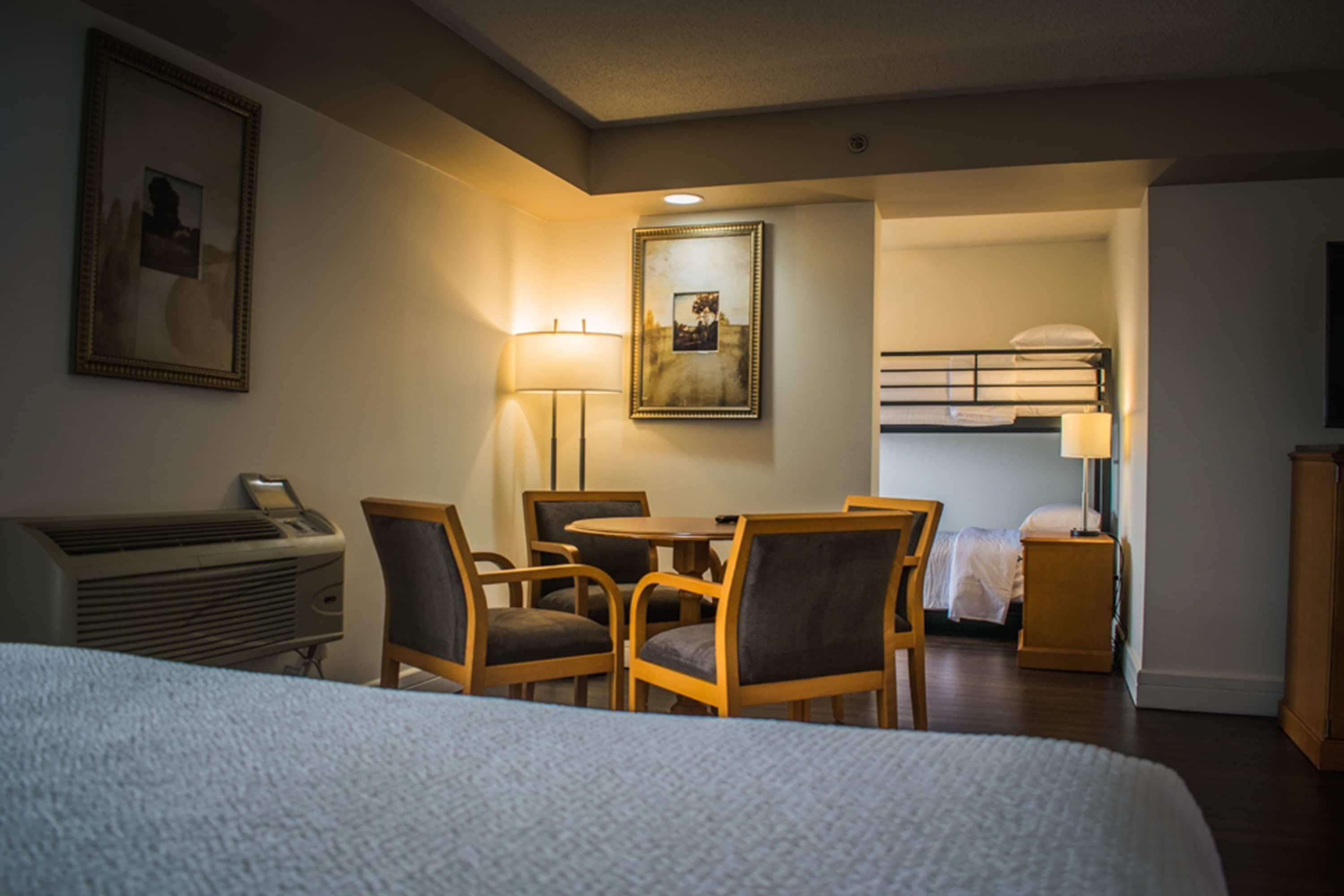 A hotel room with a bed, a desk with a chair, a table, a bunk bed, and a lamp.
