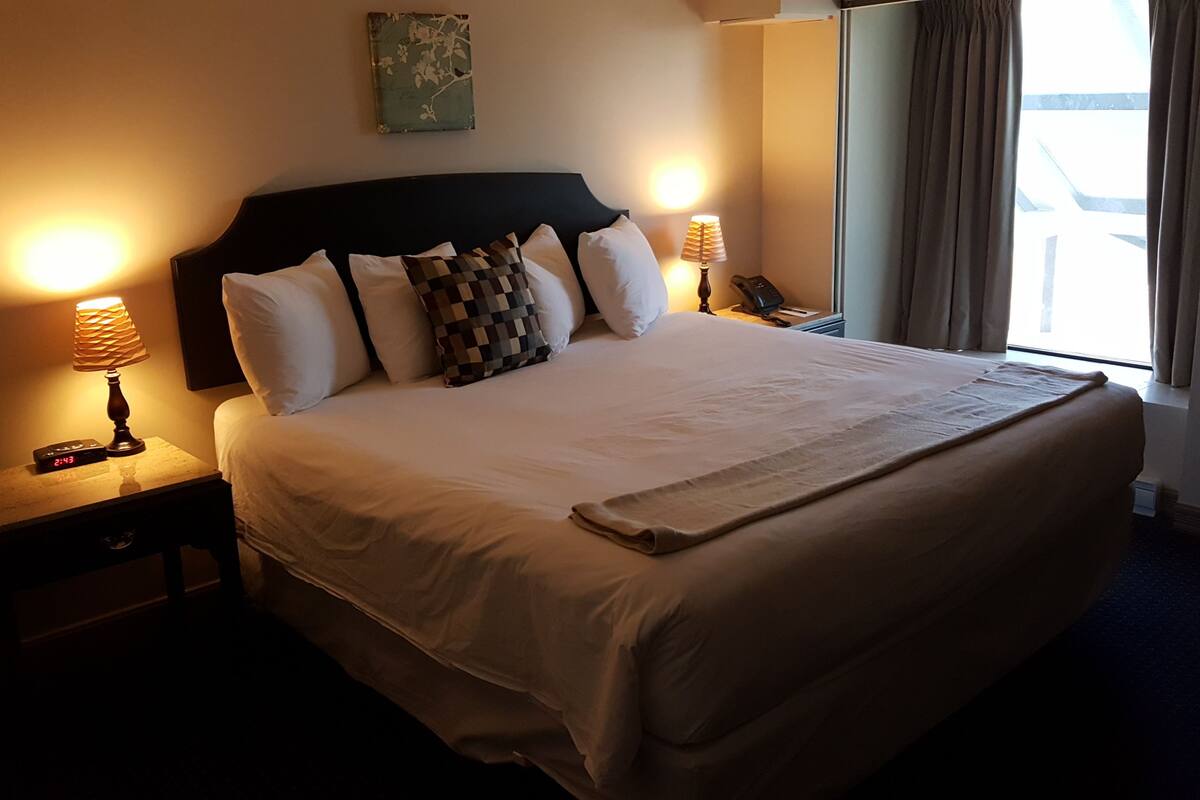 A neatly made bed with a headboard, two bedside lamps, a clock, and a window with curtains.