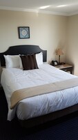 Een net opgemaakt bed met wit en bruin beddengoed, een inspirerende quote in een lijst op de muur, en een nachtkastje met een lamp en een telefoon.