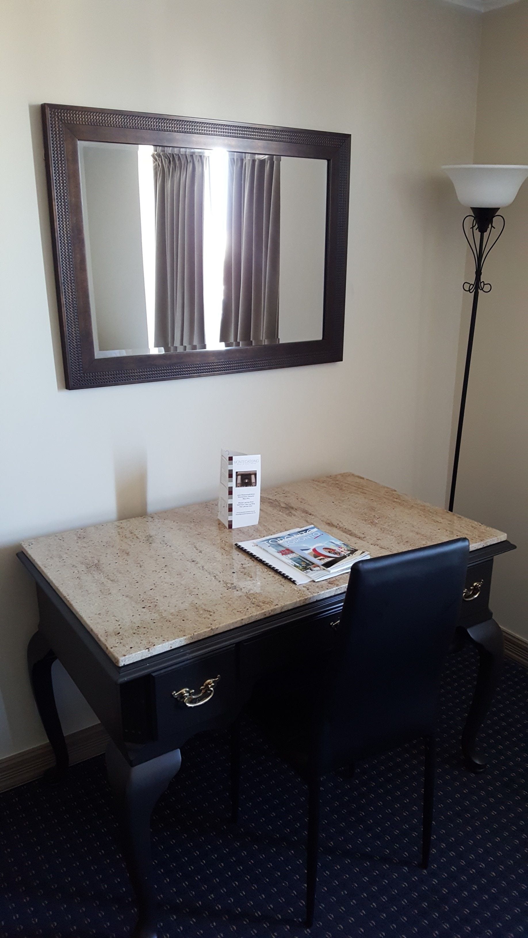 Hotelkamer met een bureau, twee stoelen, een spiegel, een lamp en een bijzettafeltje met een tijdschrift.