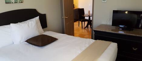 A hotel room with a bed, a TV on a dresser, and a door leading to a dining area.
