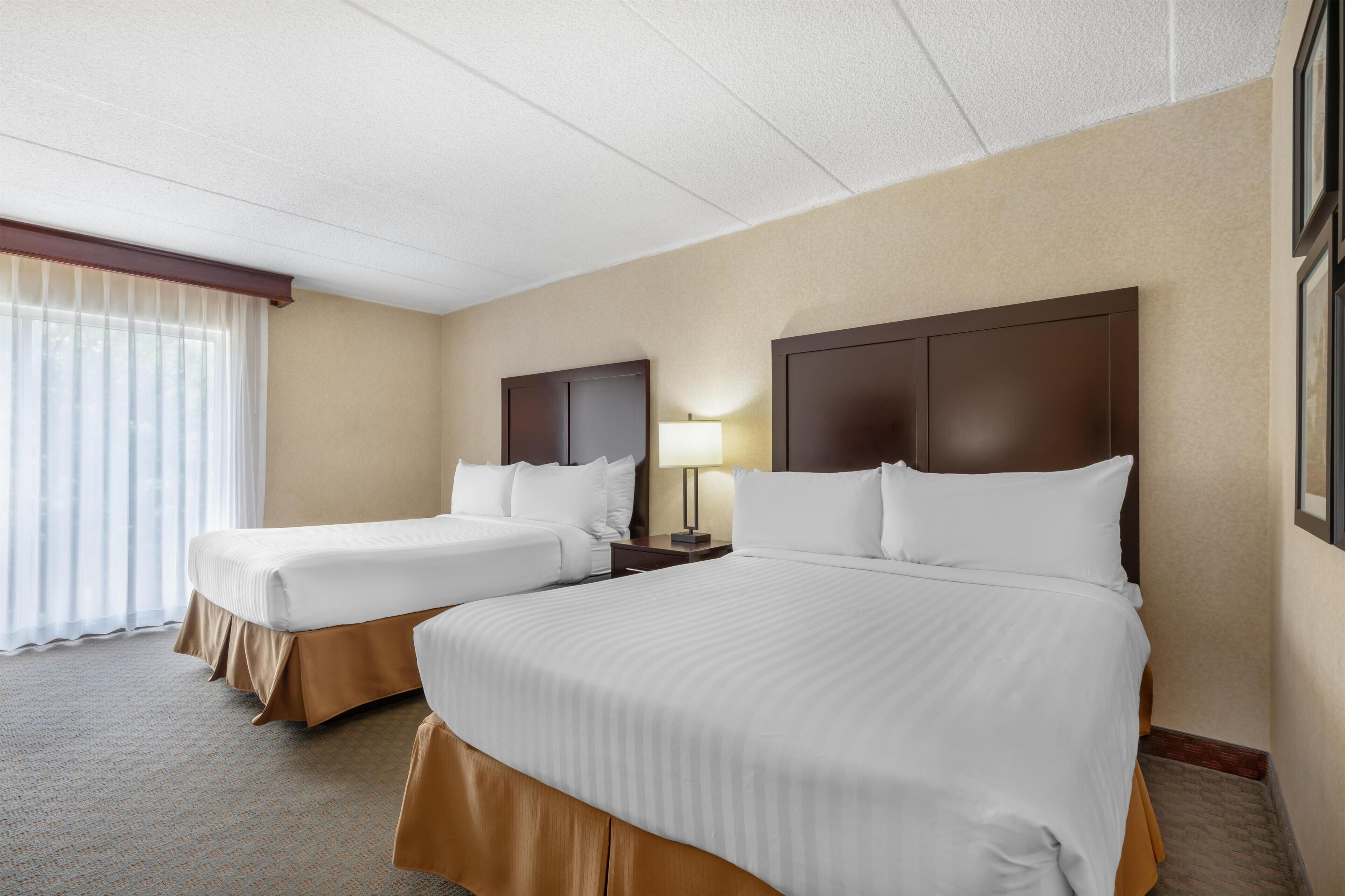Quarto de hotel com duas camas, uma mesinha de cabeceira, uma luminária e uma janela com cortinas.