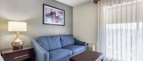 A hotel room with a sofa, a small table, a lamp, a painting on the wall, and a window with sheer curtains.