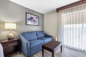 Habitación de hotel con sofá, una mesita, una lámpara, un cuadro en la pared y una ventana con cortinas traslúcidas.