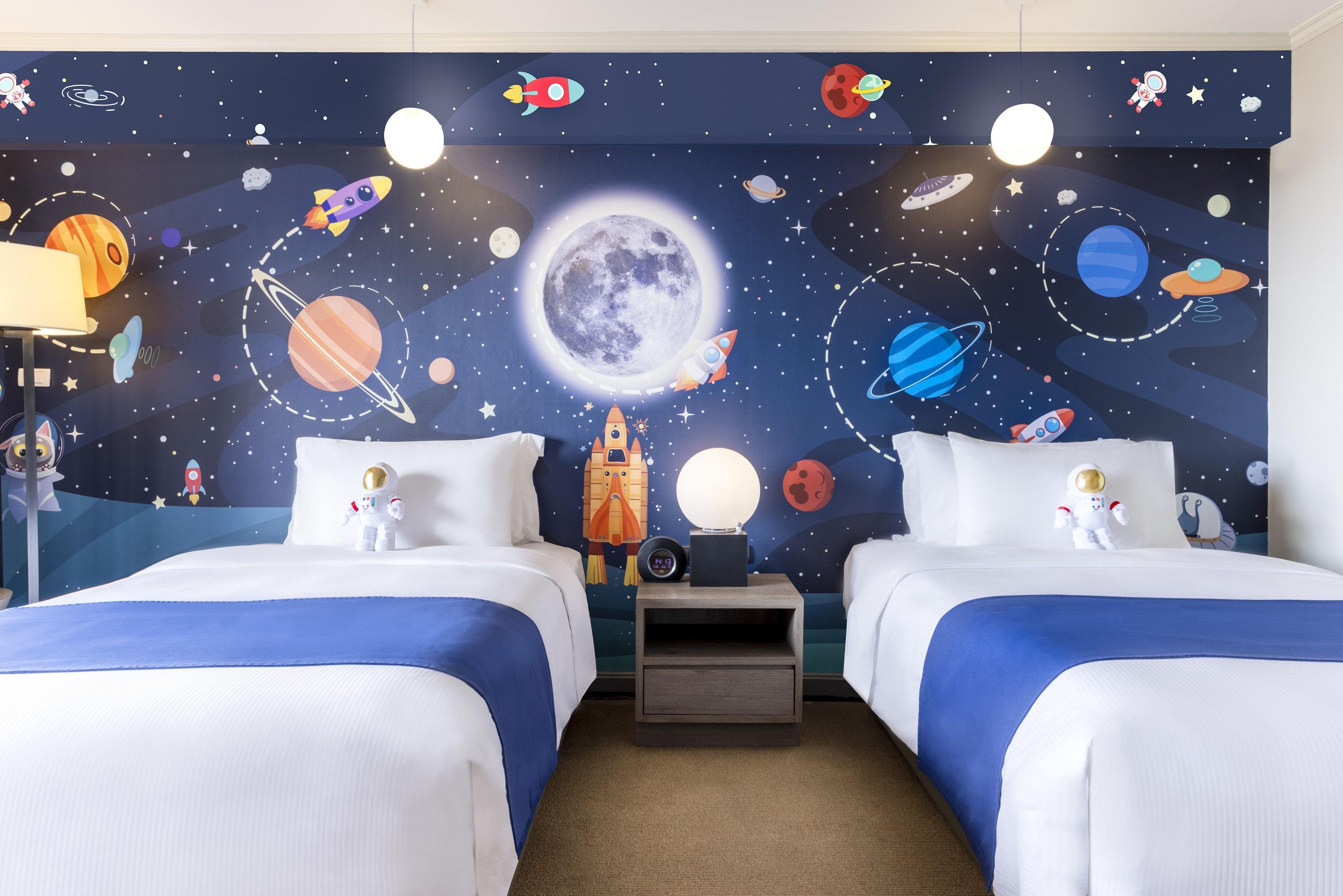 A hotel room with two beds, a nightstand, and a wall mural of space-themed decorations including planets, rockets, and an astronaut.