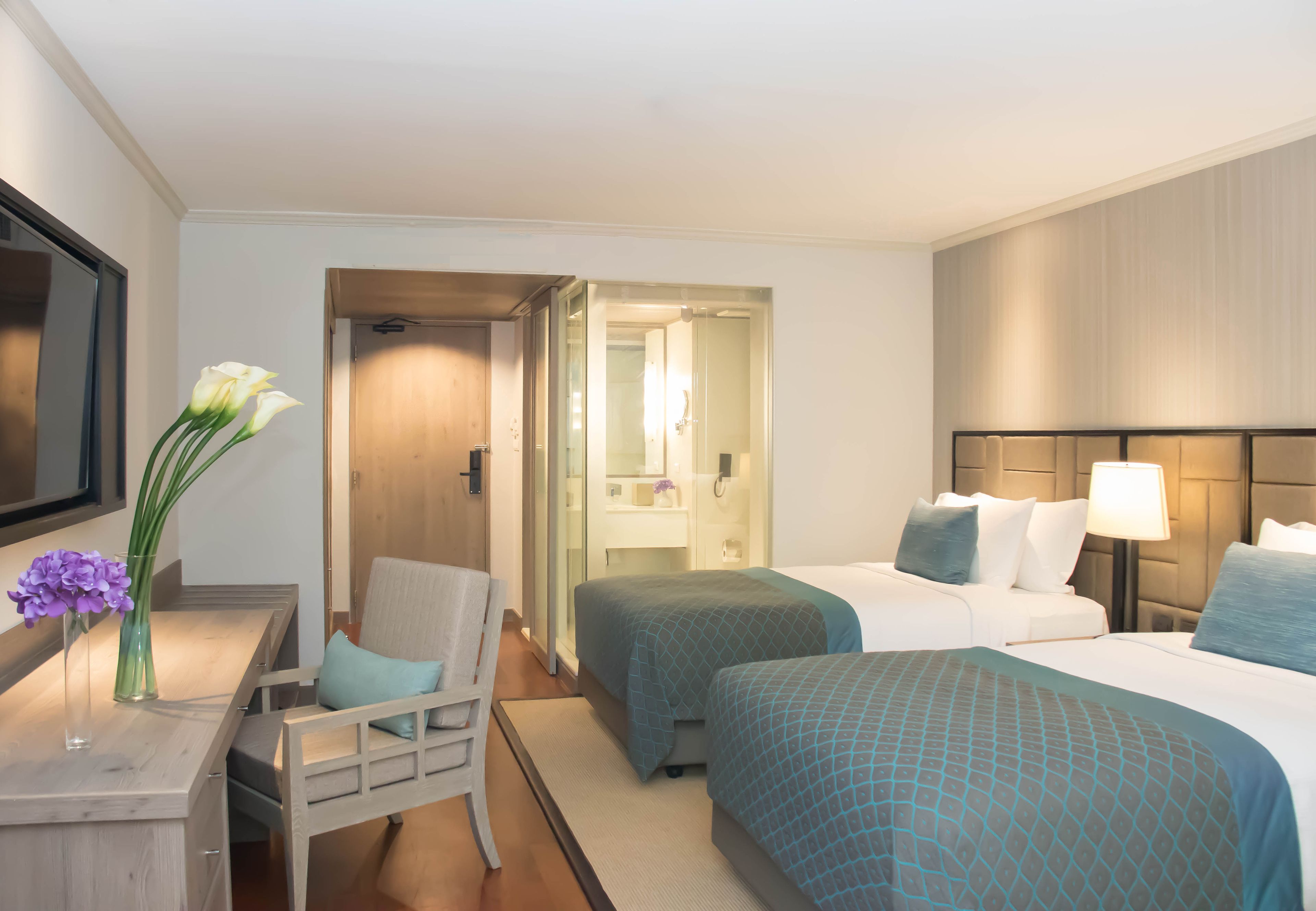 A hotel room with two beds, a desk with a chair, a mirror, and a vase with flowers.