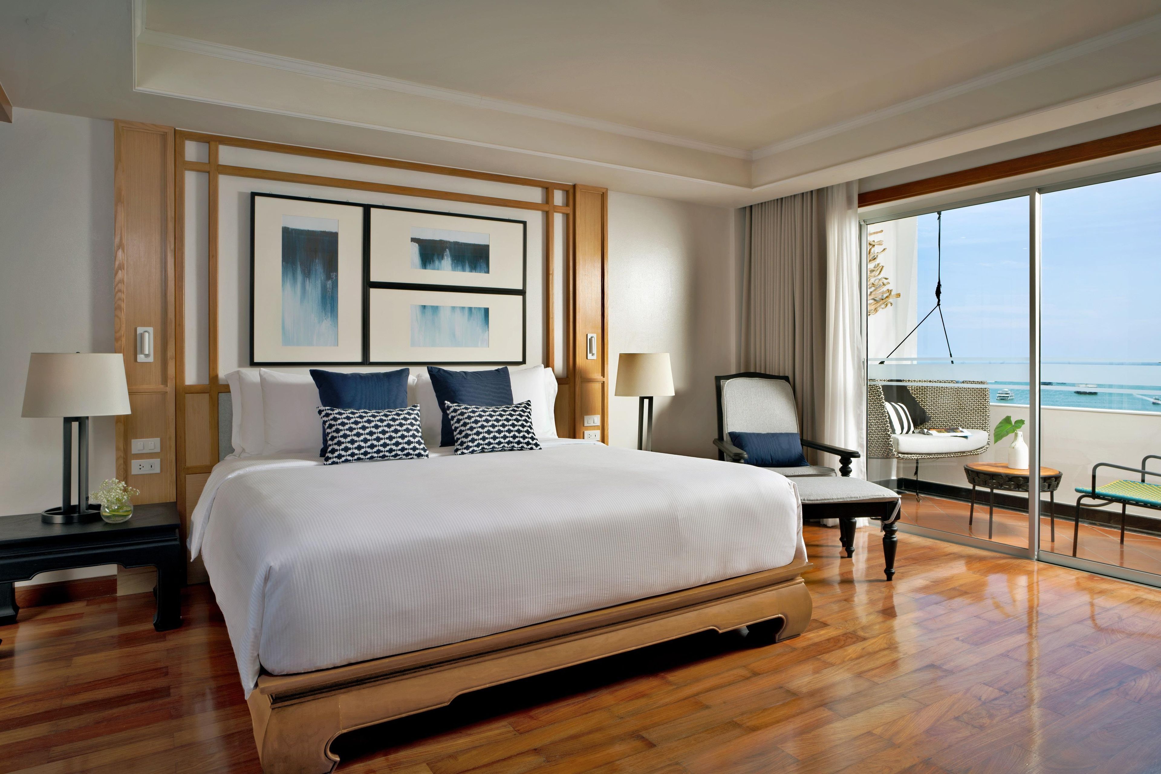 Ein Hotelzimmer mit einem großen Bett, zwei Nachttischen, einem Sessel und einem Balkon mit Meerblick, sowie Kunstwerken an der Wand.
