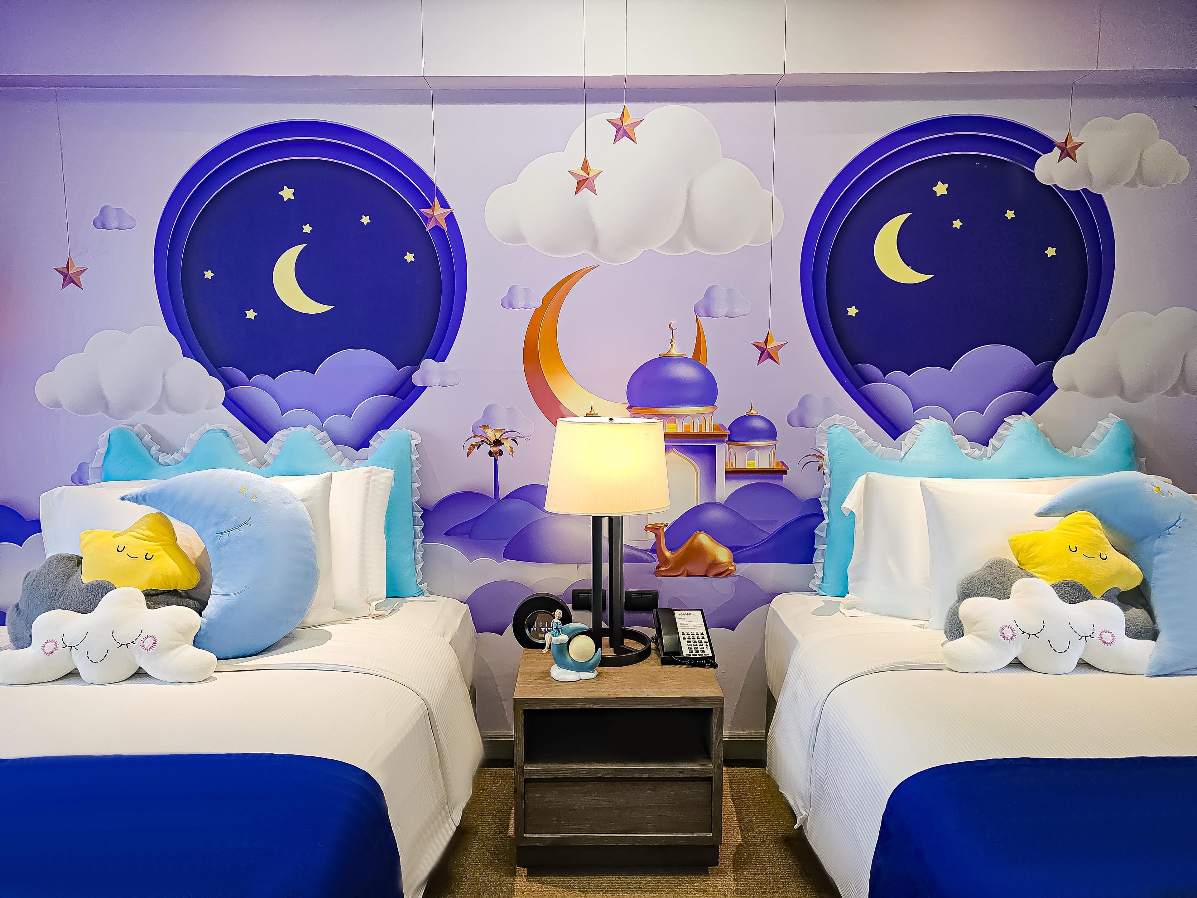 A bedroom with two beds, a nightstand, a lamp, and a wall mural of a night sky with stars and clouds.