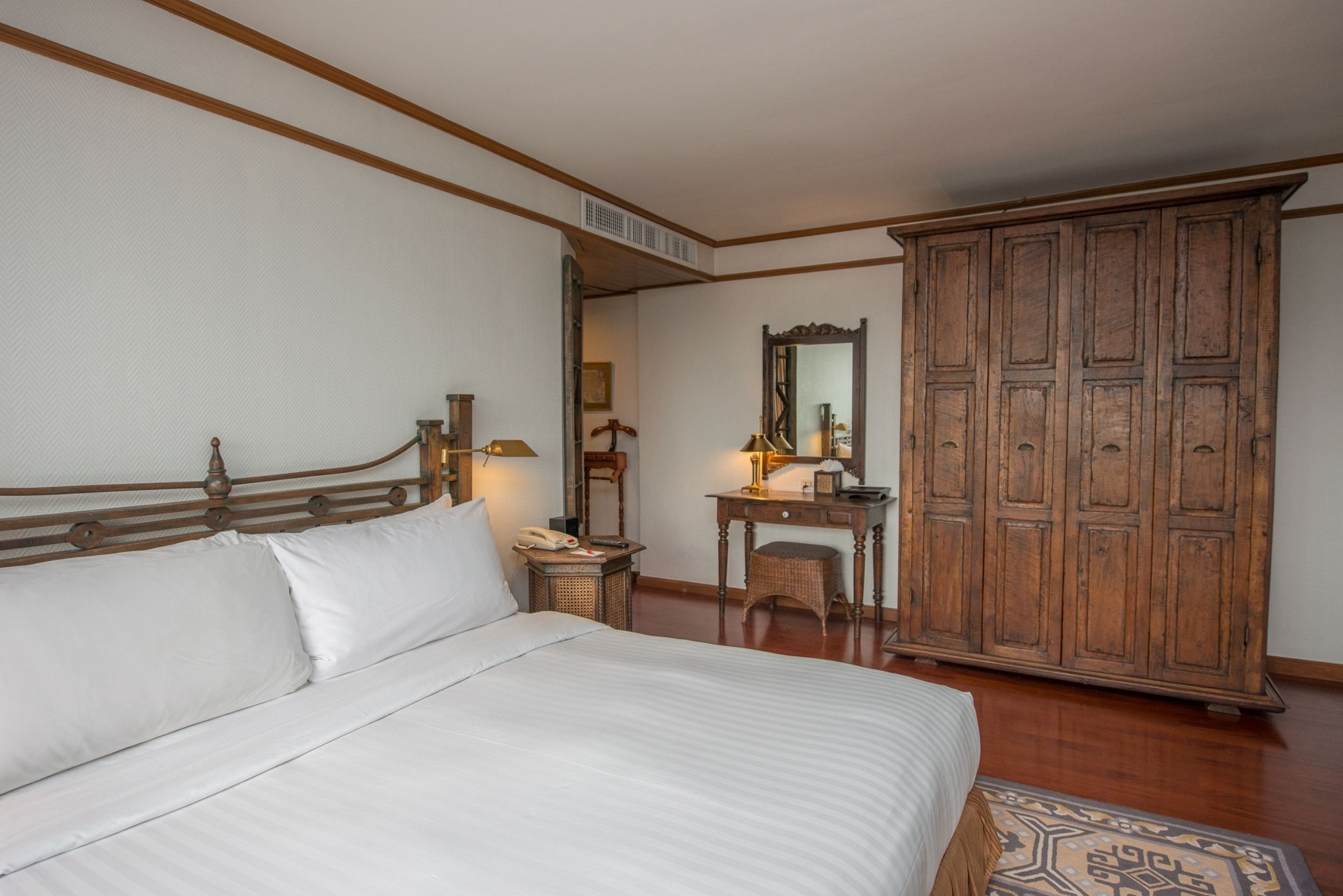A bedroom with a large bed, a wooden wardrobe, a desk with a mirror, and a wicker chair.