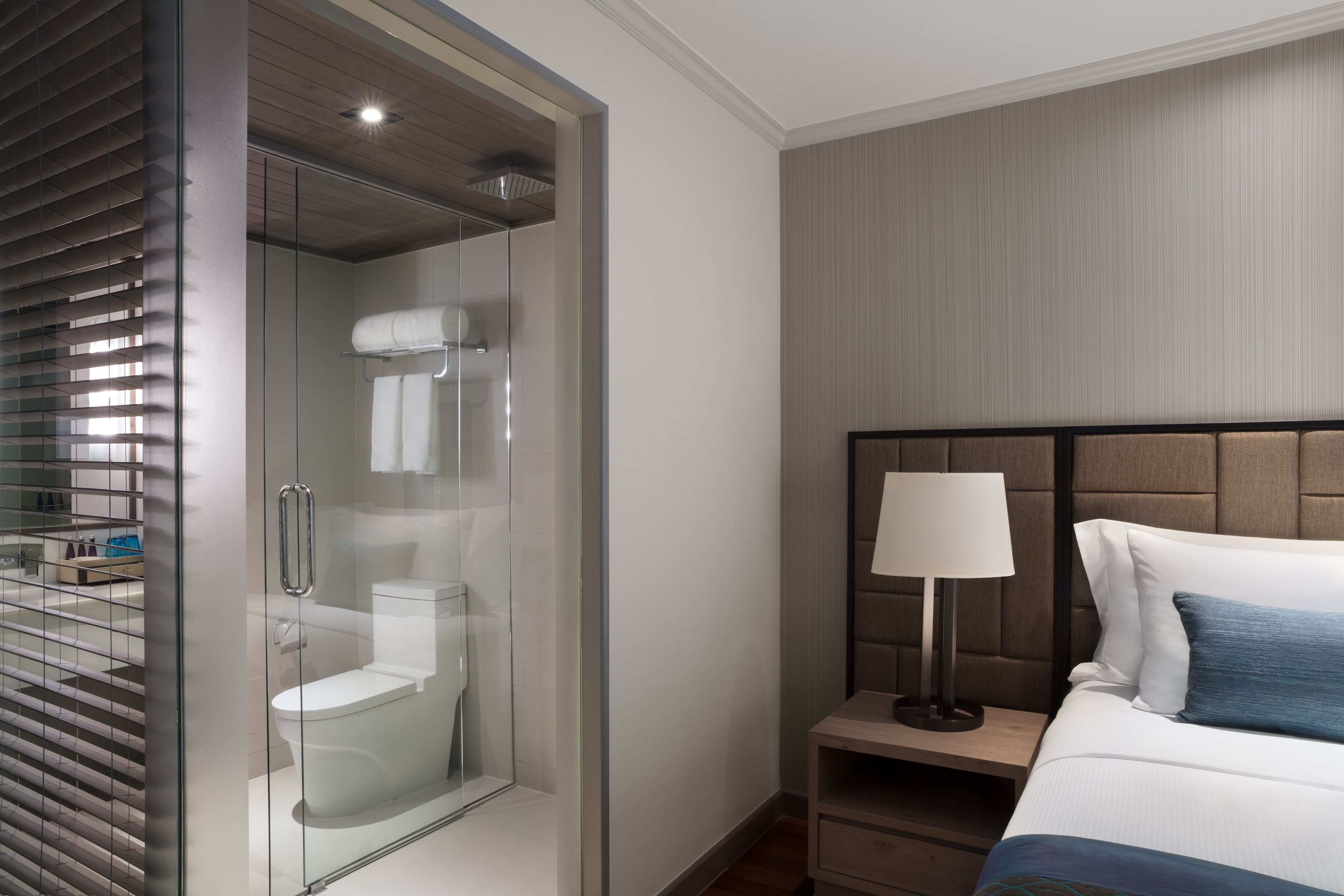 Ein modernes Hotelzimmer mit einer gläsernen Duschkabine, einem Bett mit Kopfteil, einem Nachttisch mit Lampe und einem zusätzlichen Tisch.