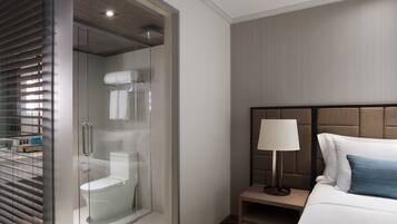 A modern hotel room with a glass-enclosed shower, a bed with a headboard, a bedside table with a lamp, and a nightstand.