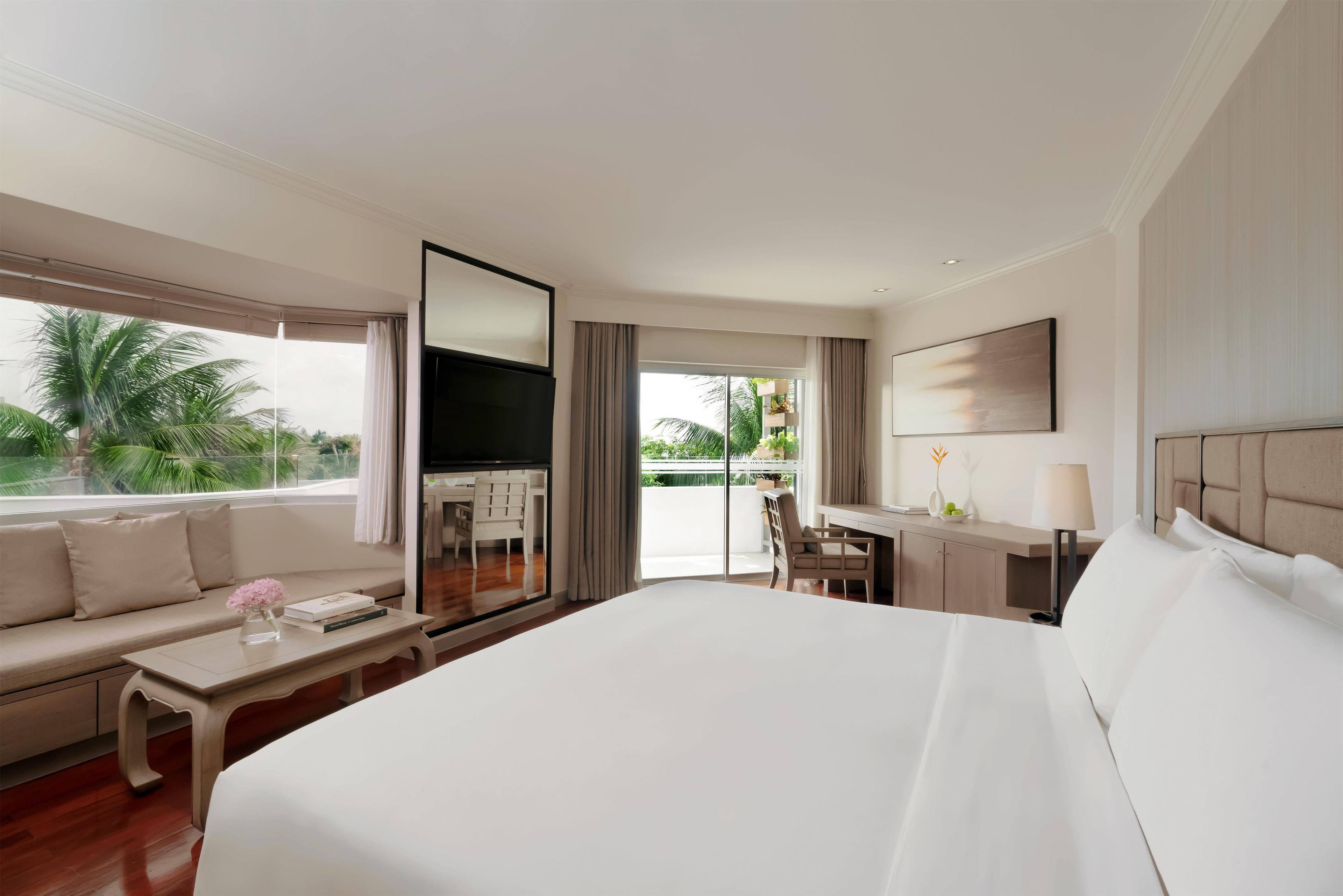 A hotel room with a large bed, a sofa, a desk, a TV, and a balcony with a view of palm trees.