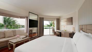 A hotel room with a large bed, a sofa, a desk, a TV, and a balcony with a view of palm trees.
