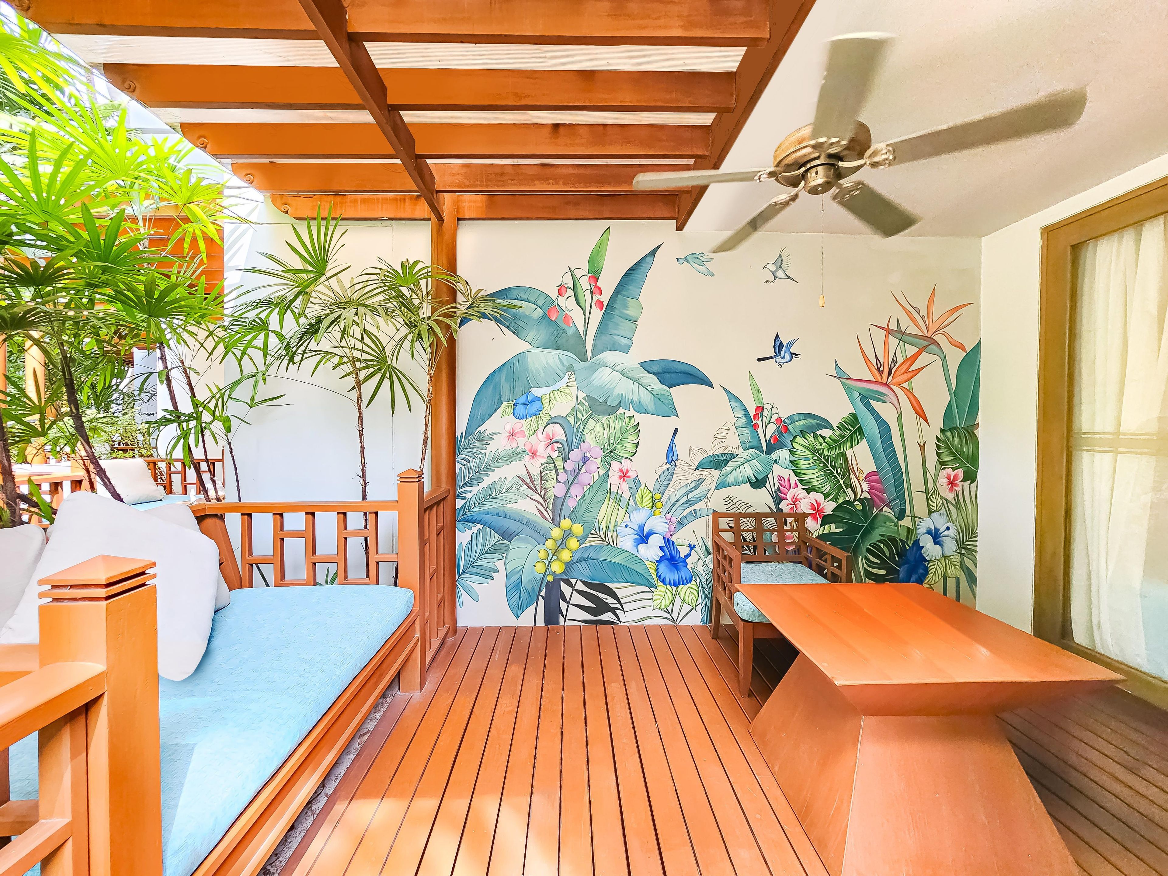 Un terrazzo in legno con una ventola a soffitto, un murale raffigurante piante tropicali e un'area salotto con un divano e un tavolo.