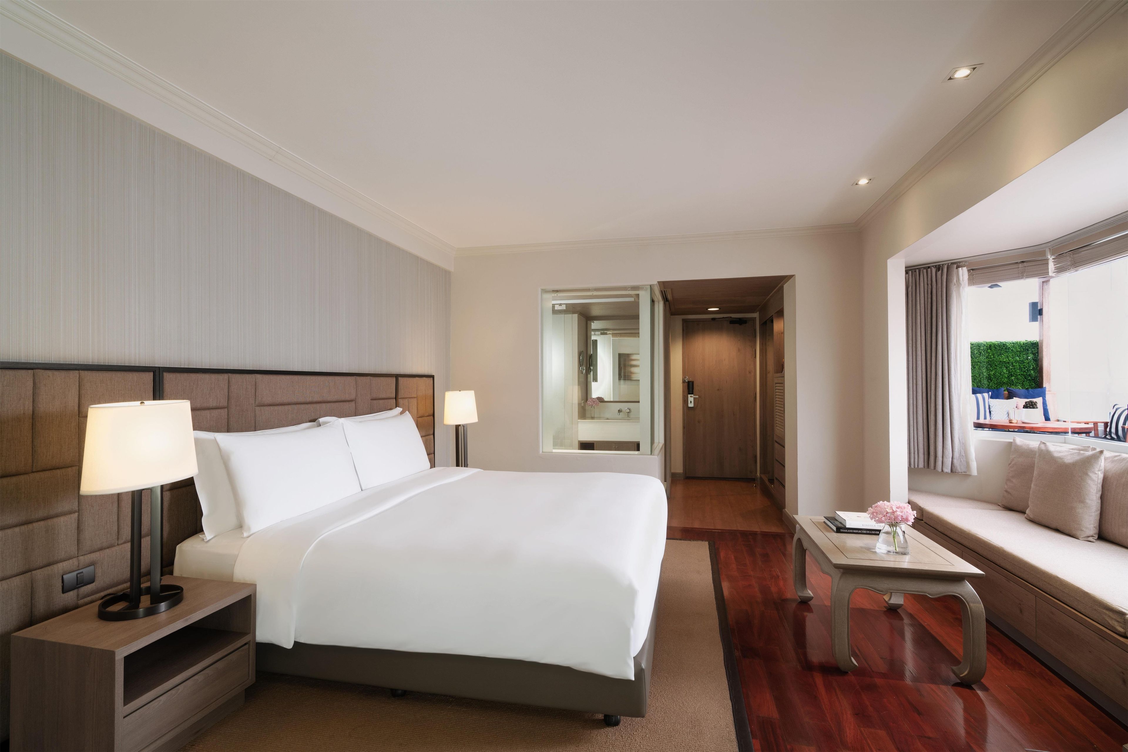 A hotel room with a large bed, bedside lamps, a sofa, a small table, and a view of the outdoors.