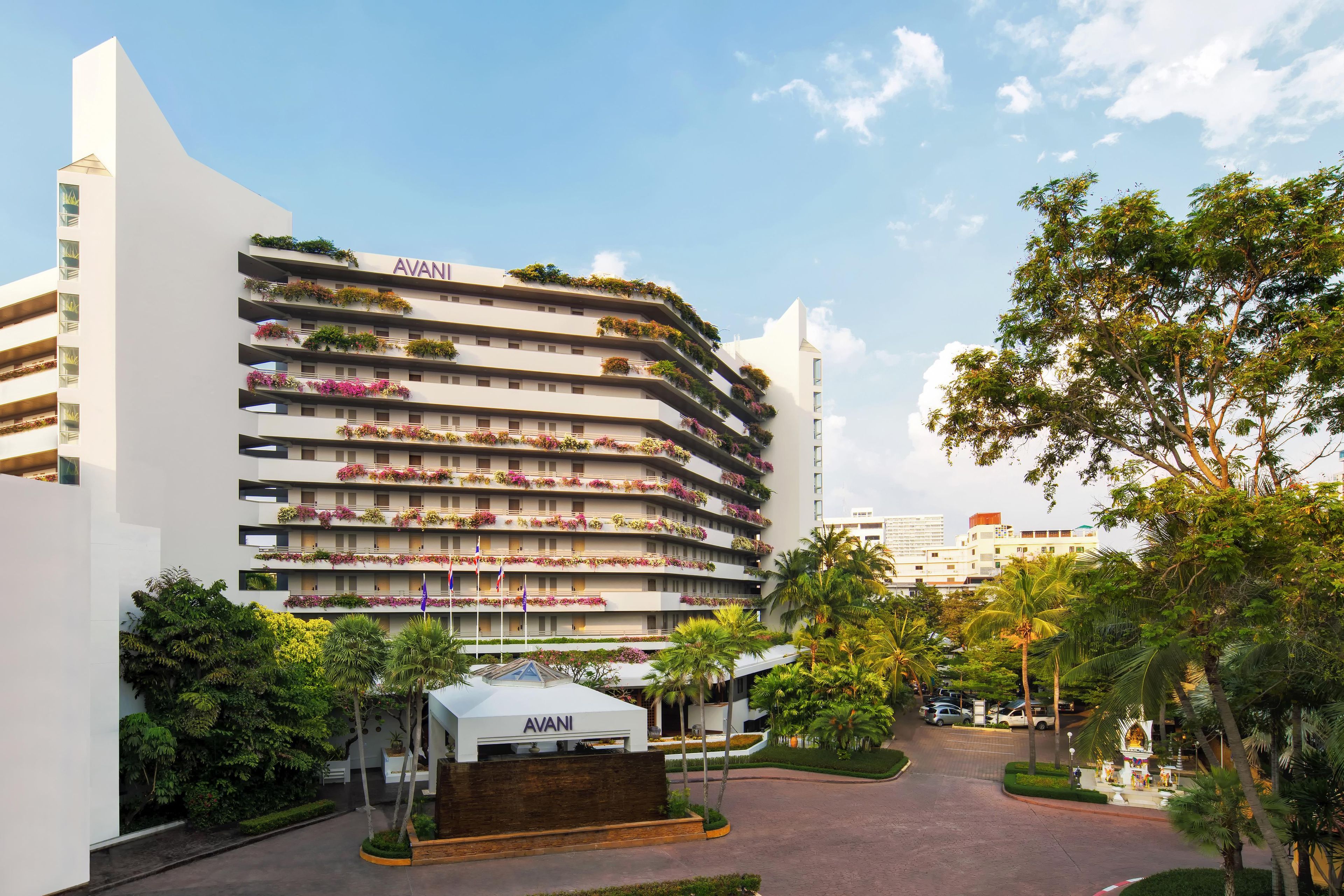 A multi-story hotel with balconies adorned with flowers, a clear sky, and a well-maintained garden area.