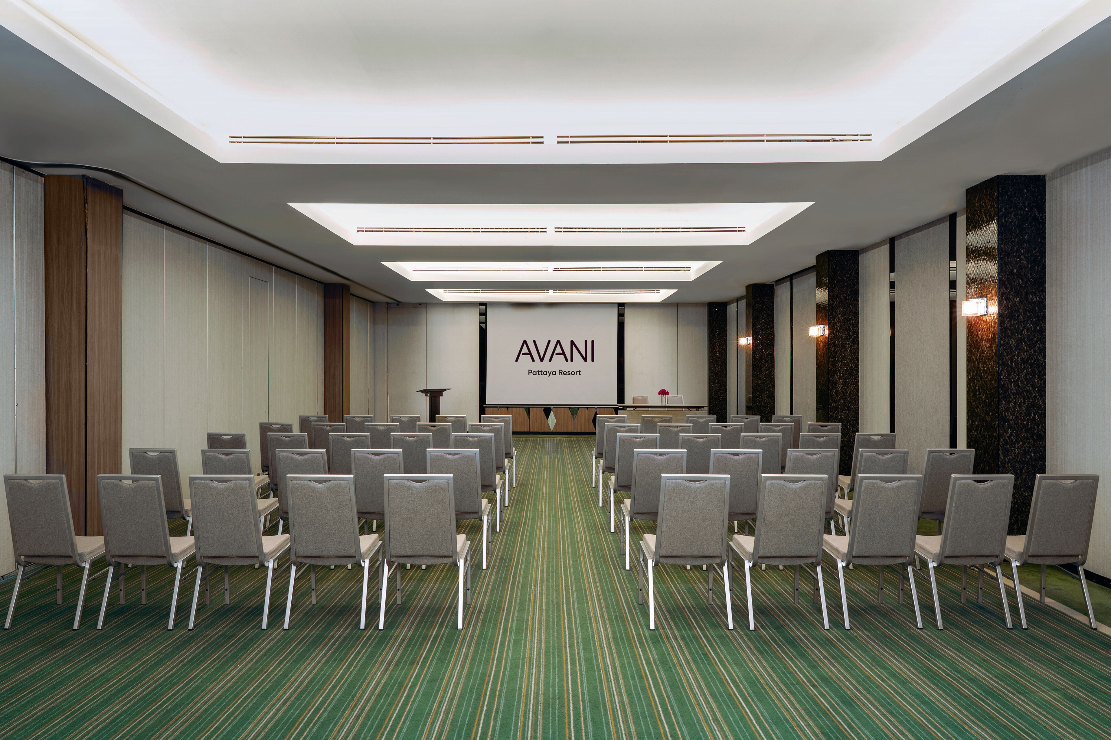 Un salón de conferencias con filas de sillas orientadas hacia una pantalla de presentación, una alfombra verde y un diseño de techo moderno.