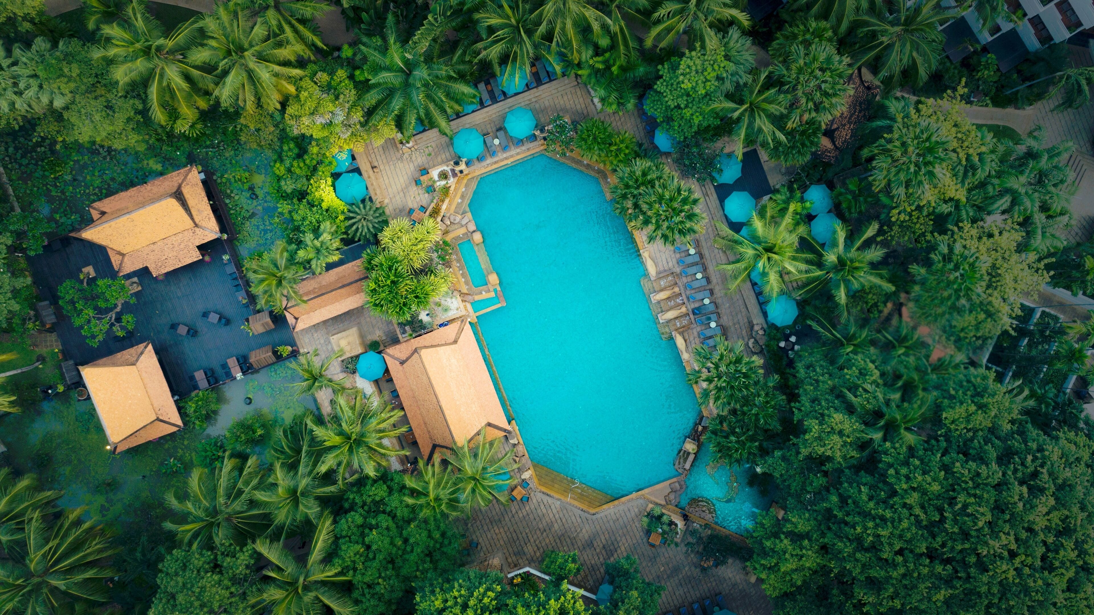 An aerial view of a resort with a central swimming pool, multiple lounge areas, and surrounding lush greenery.