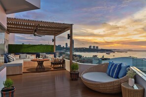 Terraço no telhado com área de estar, mesa e vista do horizonte urbano e do mar ao pôr do sol.