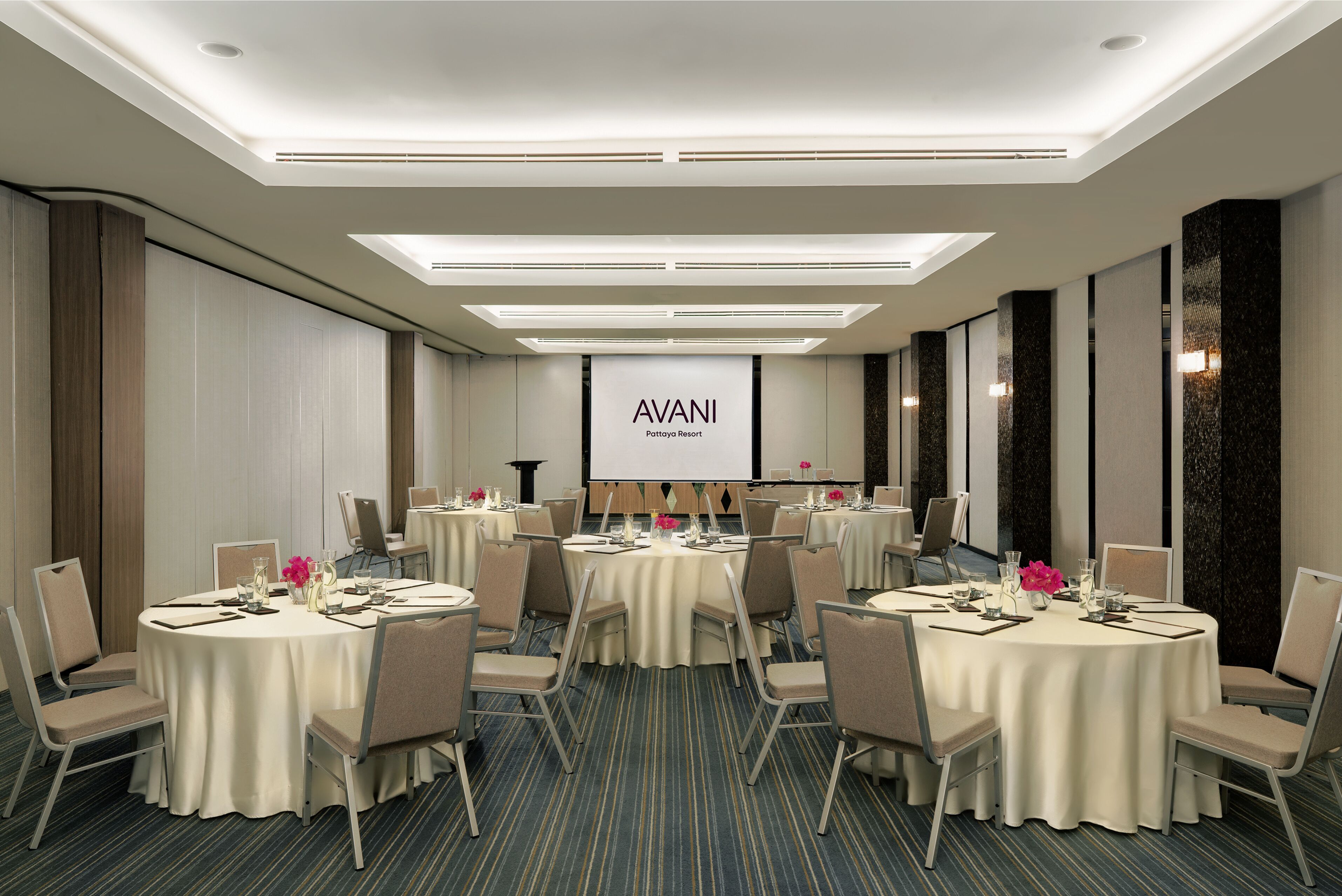 Un salón de conferencias con varias mesas redondas, cada una con sillas y preparación para comidas, con una pantalla grande al fondo.