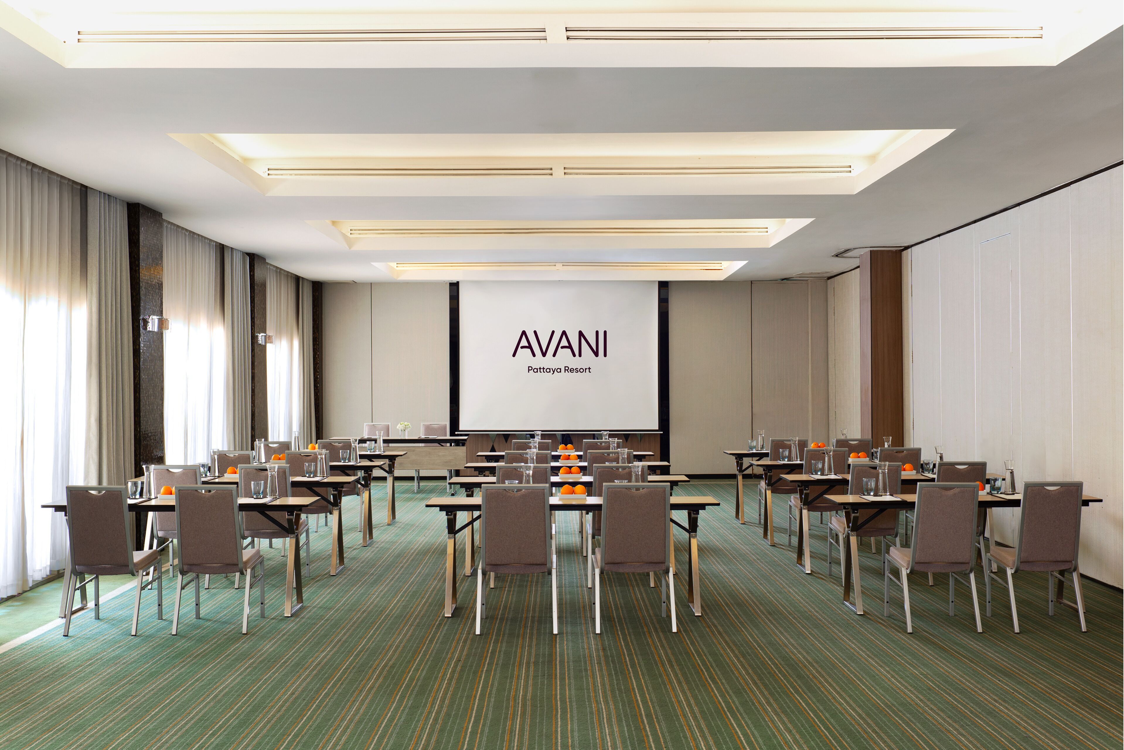 Una sala de conferencias en Pattaya Resort con la marca AVANI, con varias mesas y sillas, una pantalla grande y una alfombra verde a rayas.
