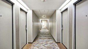 A long, narrow hallway with doors on both sides, a patterned carpet, and a ceiling-mounted fire alarm. - Hampton Inn Detroit/Madison Heights/South Troy (Madison Heights)