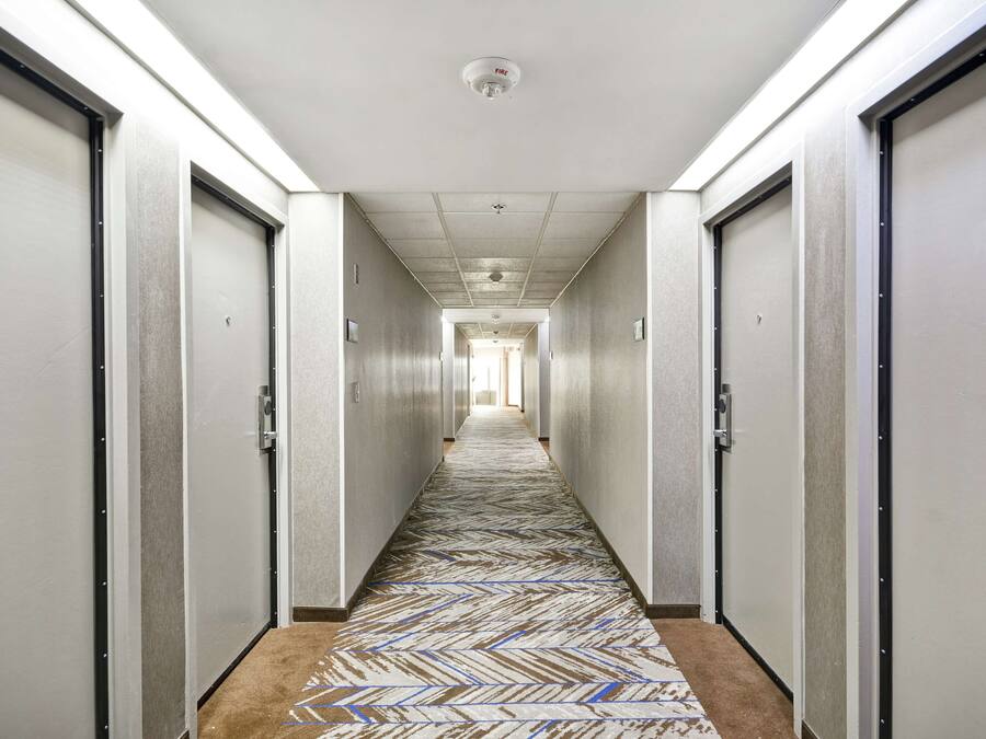 A long, narrow hallway with doors on both sides, a patterned carpet, and a ceiling-mounted fire alarm.