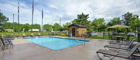 A swimming pool with lounge chairs and a small building, surrounded by trees and flags.