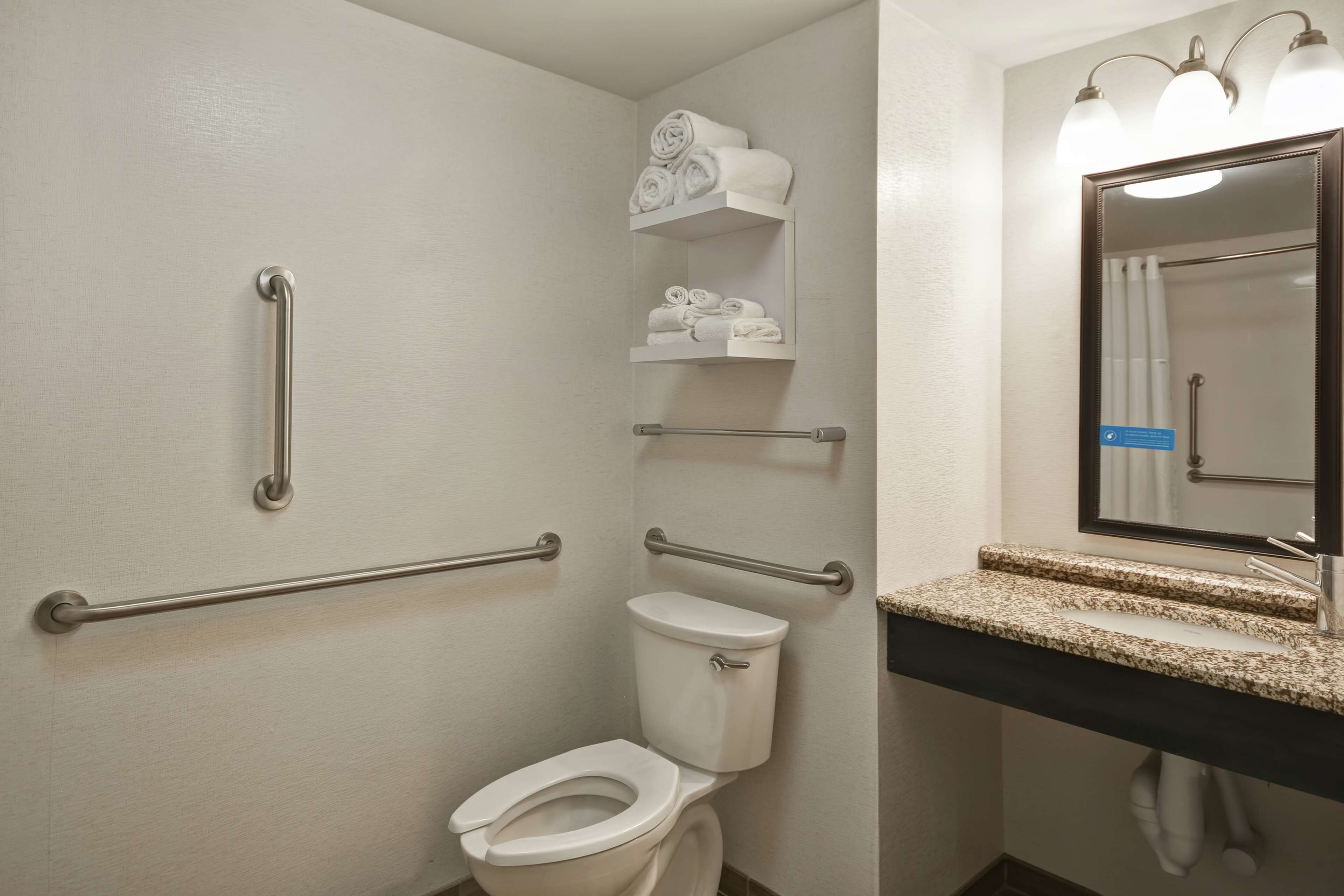 Une salle de bain équipée de toilettes, de barres d’appui, d’un lavabo avec un miroir et d’étagères pour ranger les serviettes.