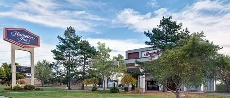 A Hampton Inn by Hilton hotel with a large sign, surrounded by trees and a grassy area.
