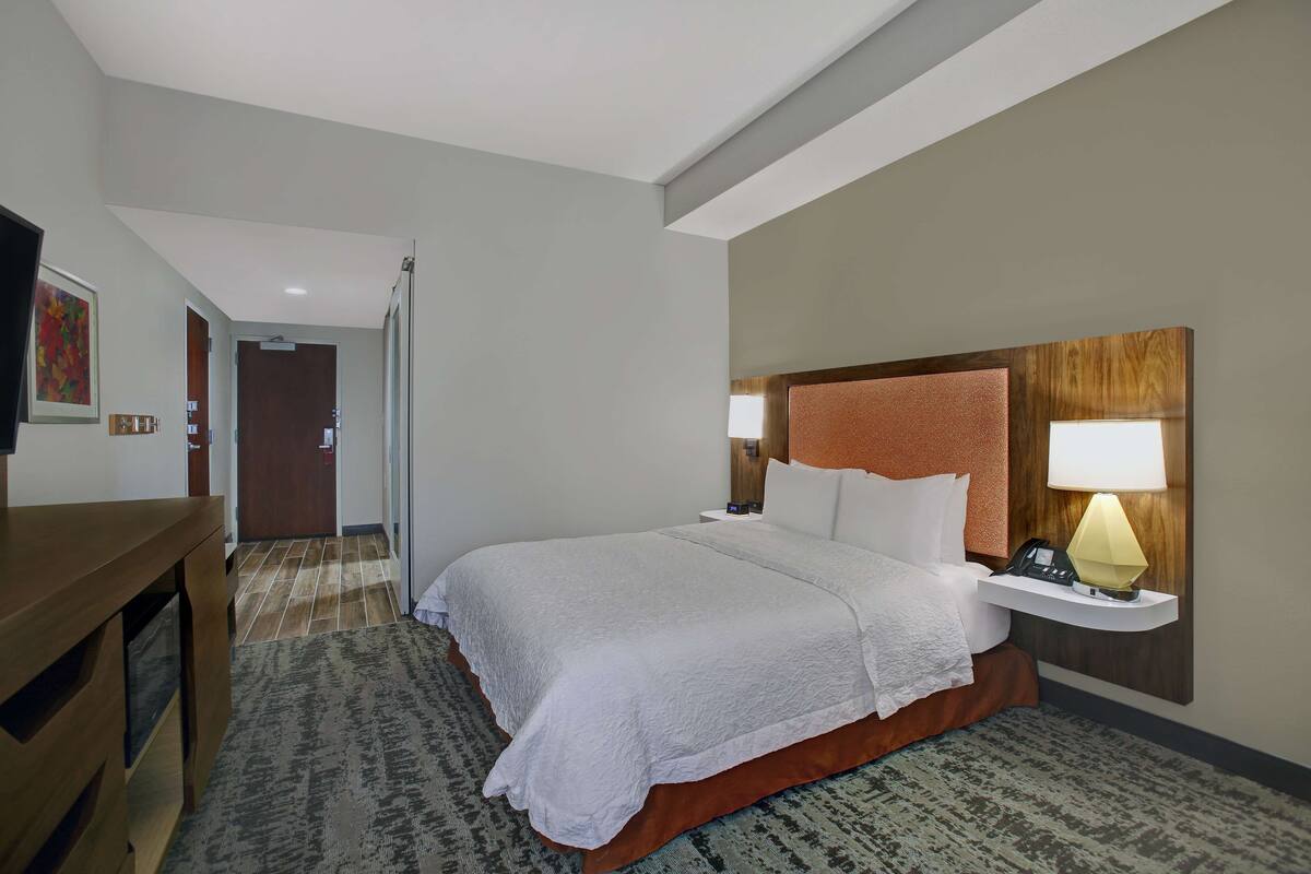 A hotel room with a large bed, a bedside table with a lamp, a TV, and a door leading to another room.