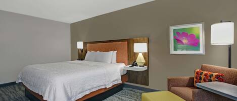 Habitación de hotel con cama, una silla, una mesita, una lámpara y un cuadro en la pared.