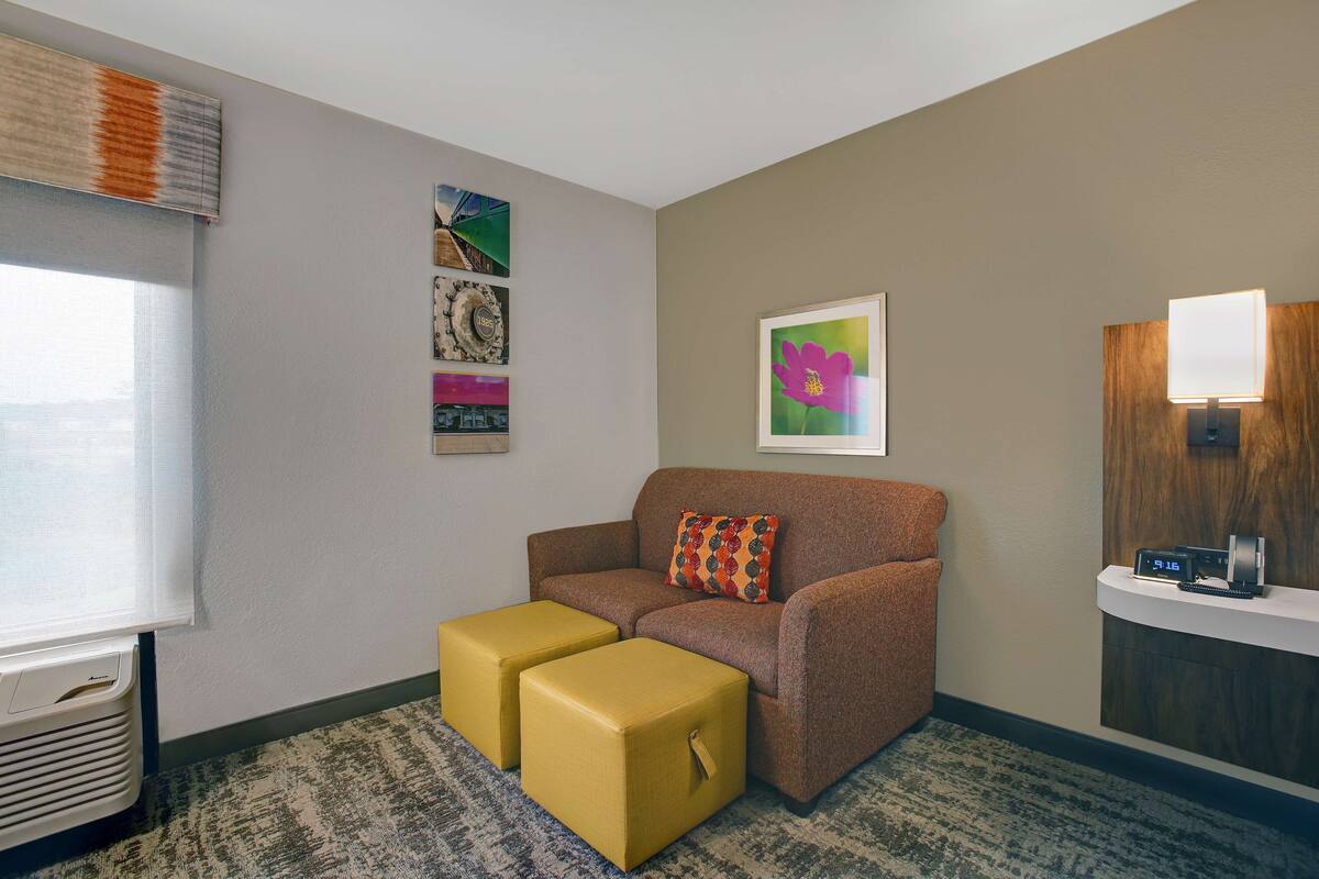 A hotel room with a brown sofa, two yellow ottomans, a window with blinds, a wall-mounted clock, and framed artwork.