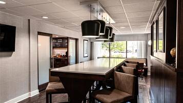 Un intérieur de restaurant moderne avec une longue table à manger, de nombreuses chaises, des luminaires suspendus et une vue sur l’extérieur.