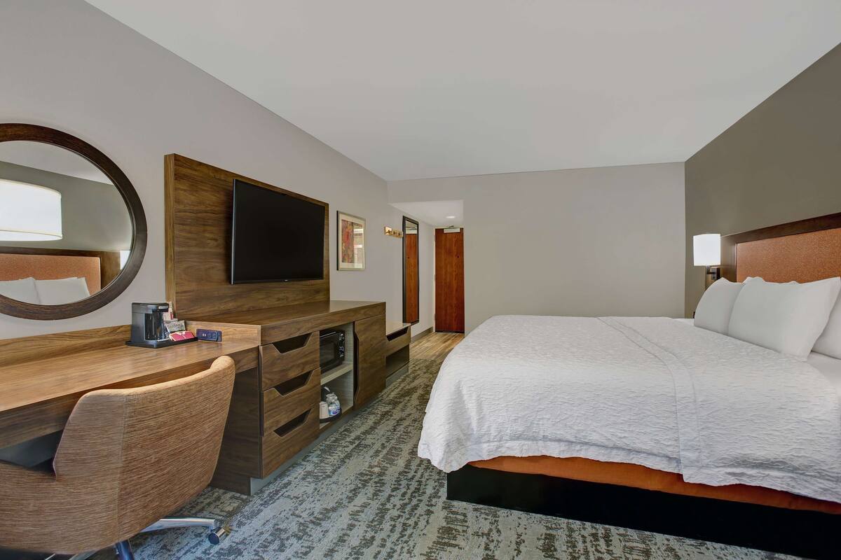 A hotel room with a bed, a desk with a chair, a television, a mirror, and a door.