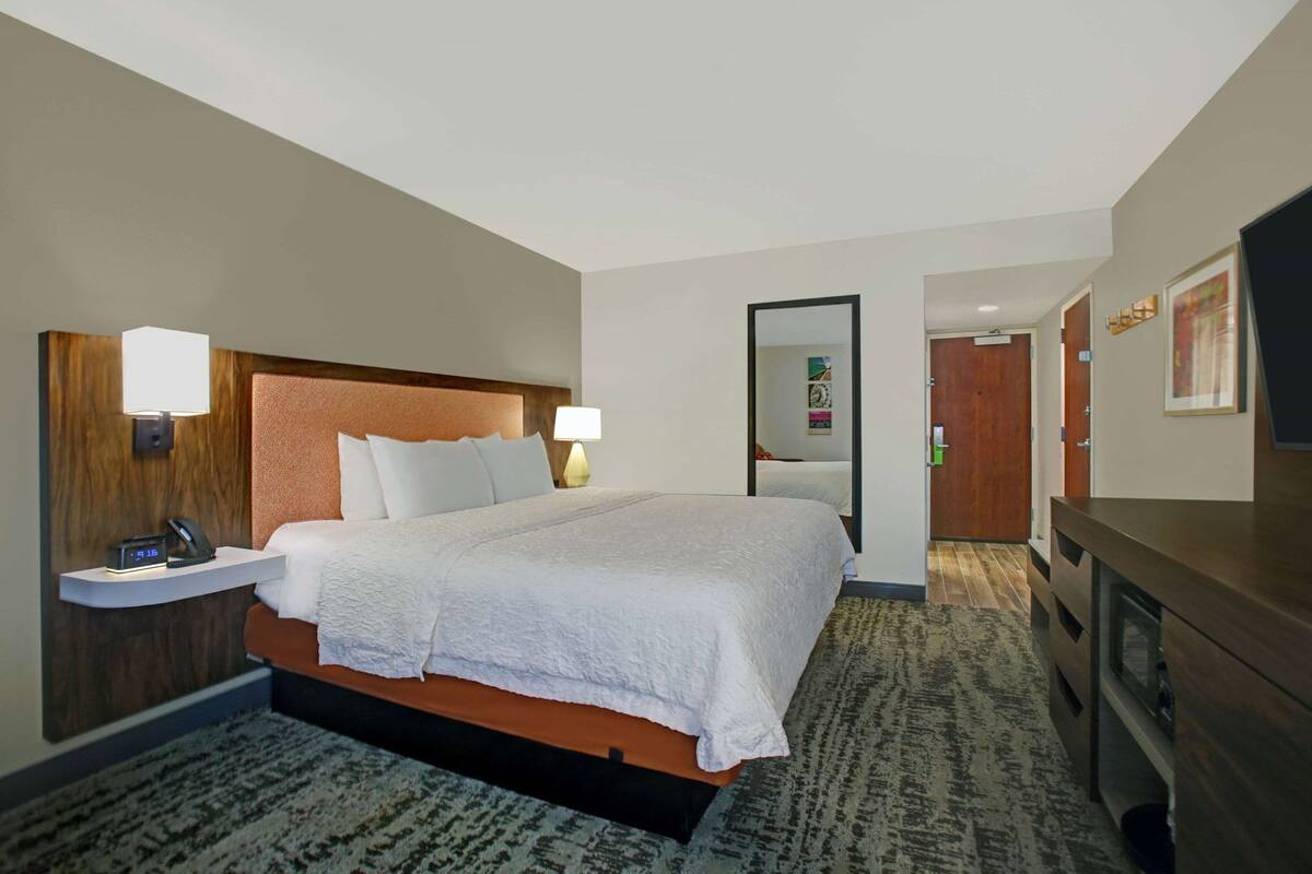 A hotel room with a bed, bedside table with a lamp, a clock, a mirror, a dresser, and a door.