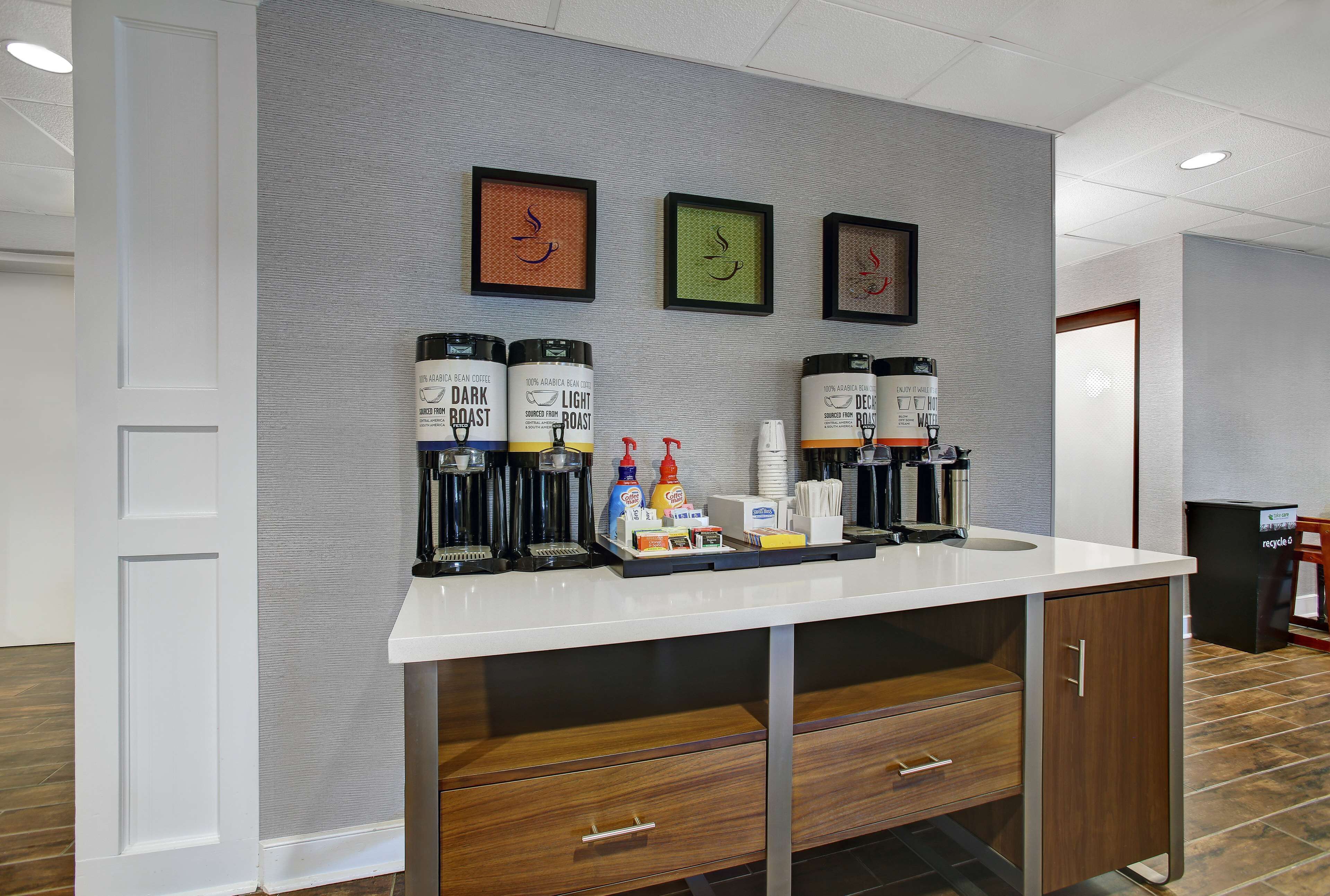 A clean, modern kitchenette with a coffee station featuring three coffee makers, condiment dispensers, and a small tray.