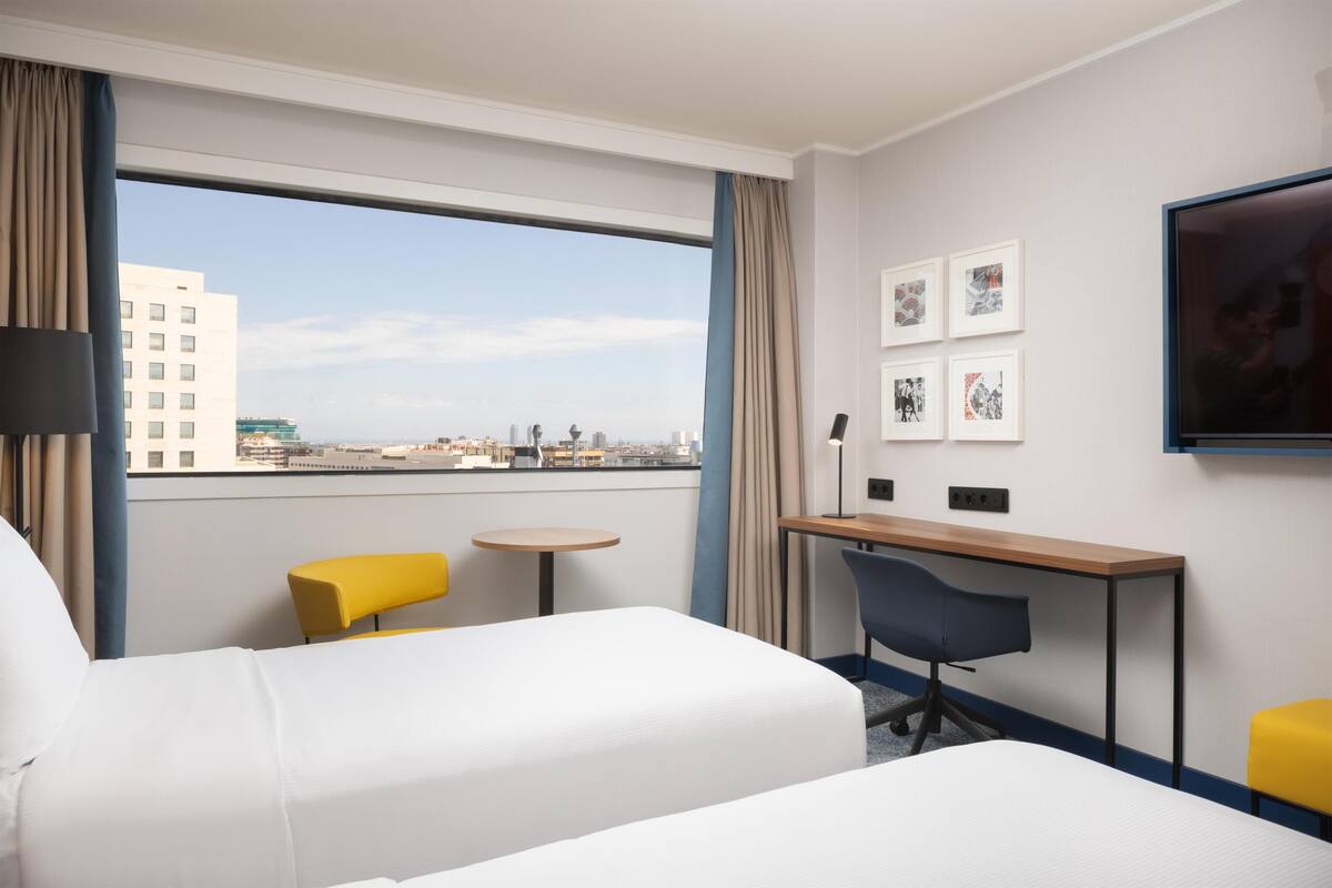 A hotel room with a bed, a desk, two chairs, a TV, and a window with a city view.